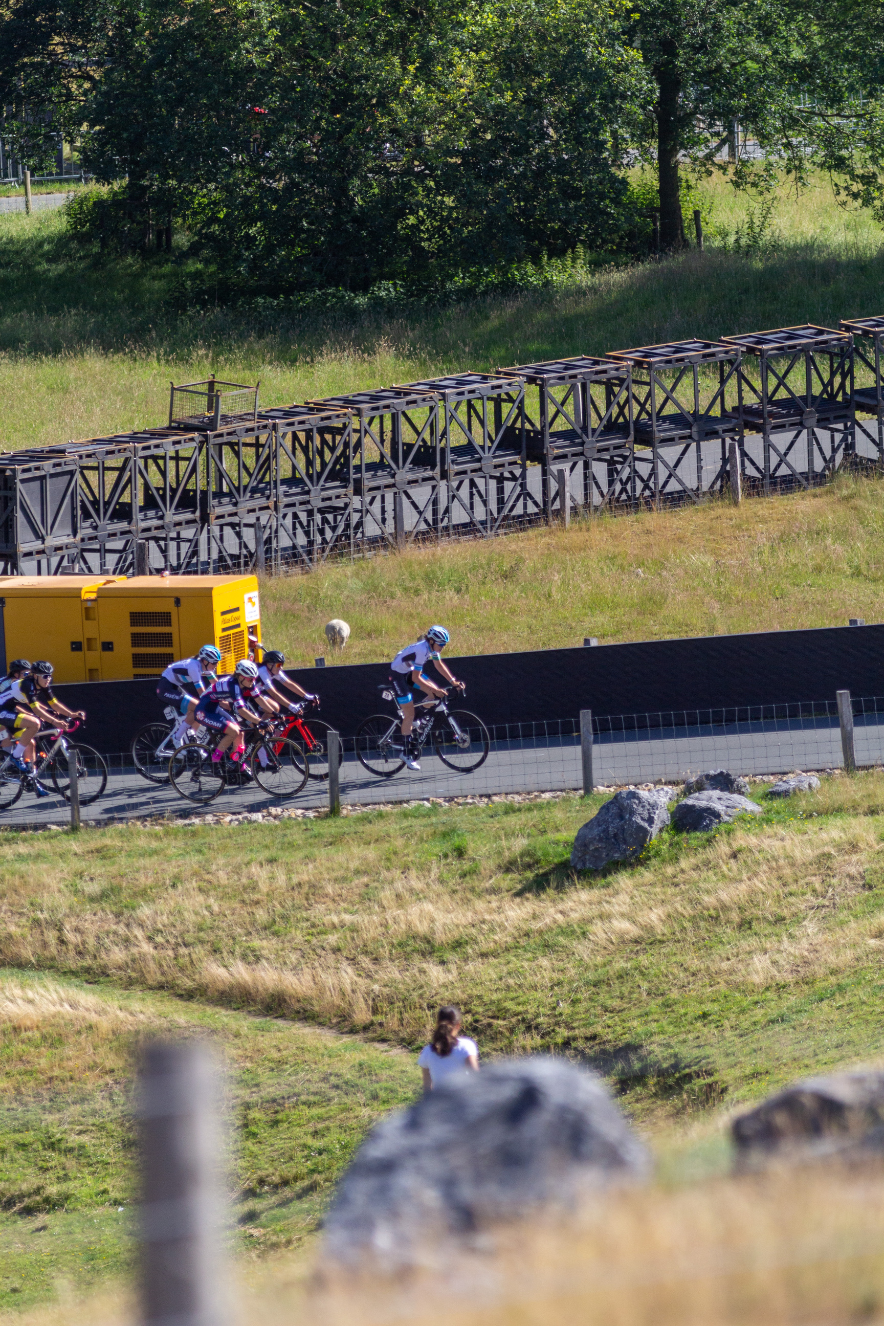 A group of cyclists in a race on a road with a large structure behind them.