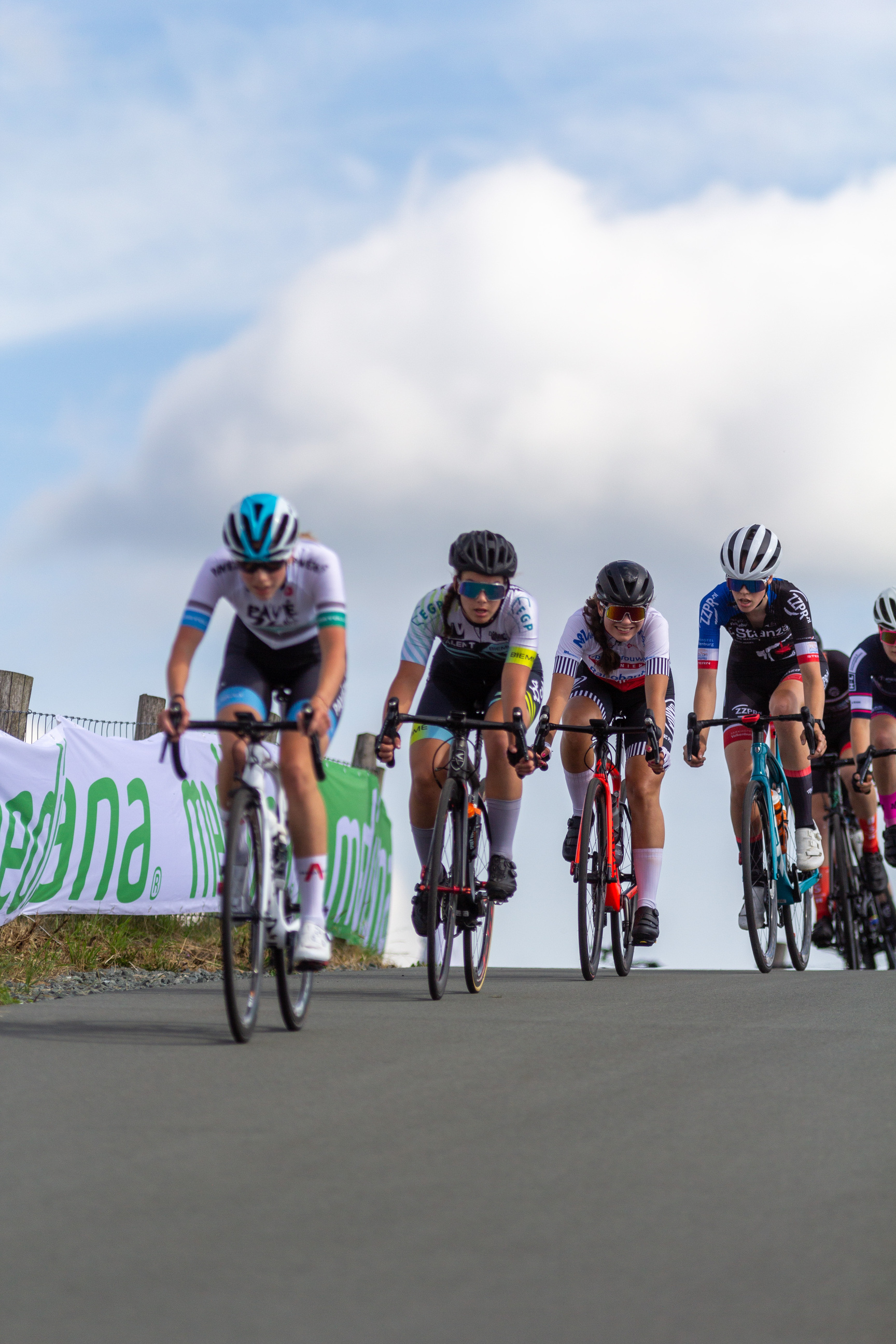 A group of bicyclists, sponsored by NK Dames, compete on a road during Wielrennen.