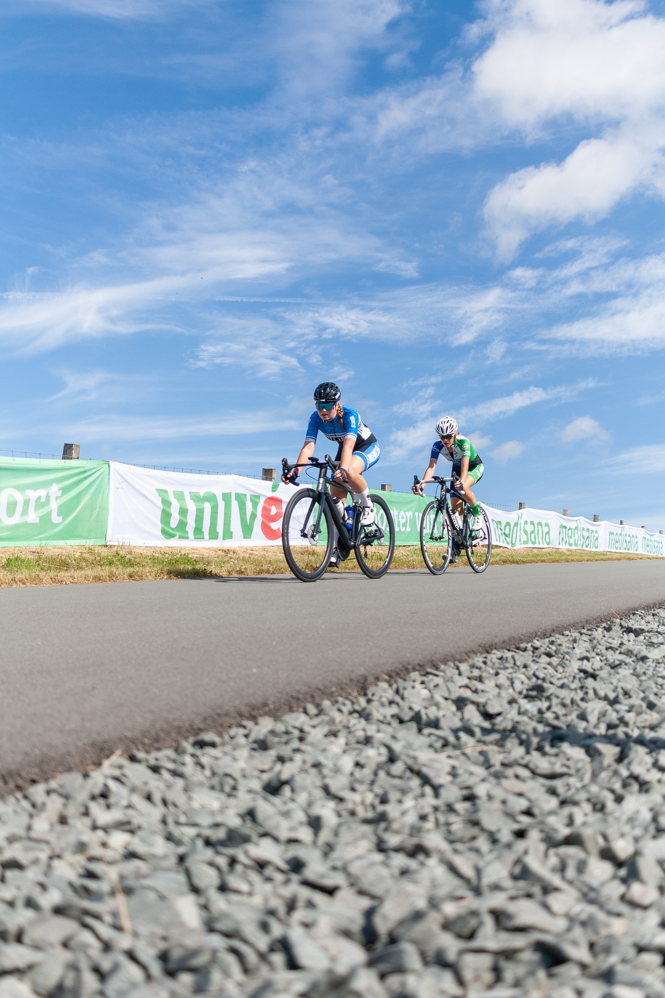 Two cyclists ride side by side on a gravel road with a banner displaying "University Sport" in the background.