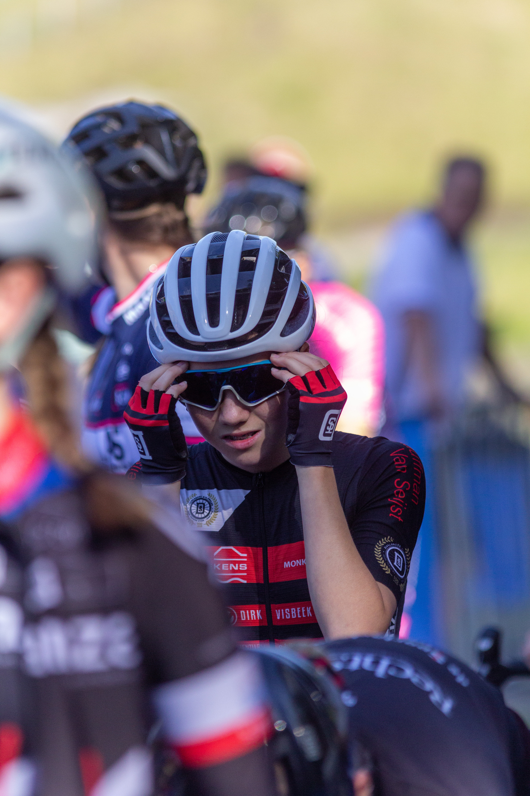 A group of cyclists with a red and black jersey on that says "NK Dames" are wearing helmets and sunglasses.