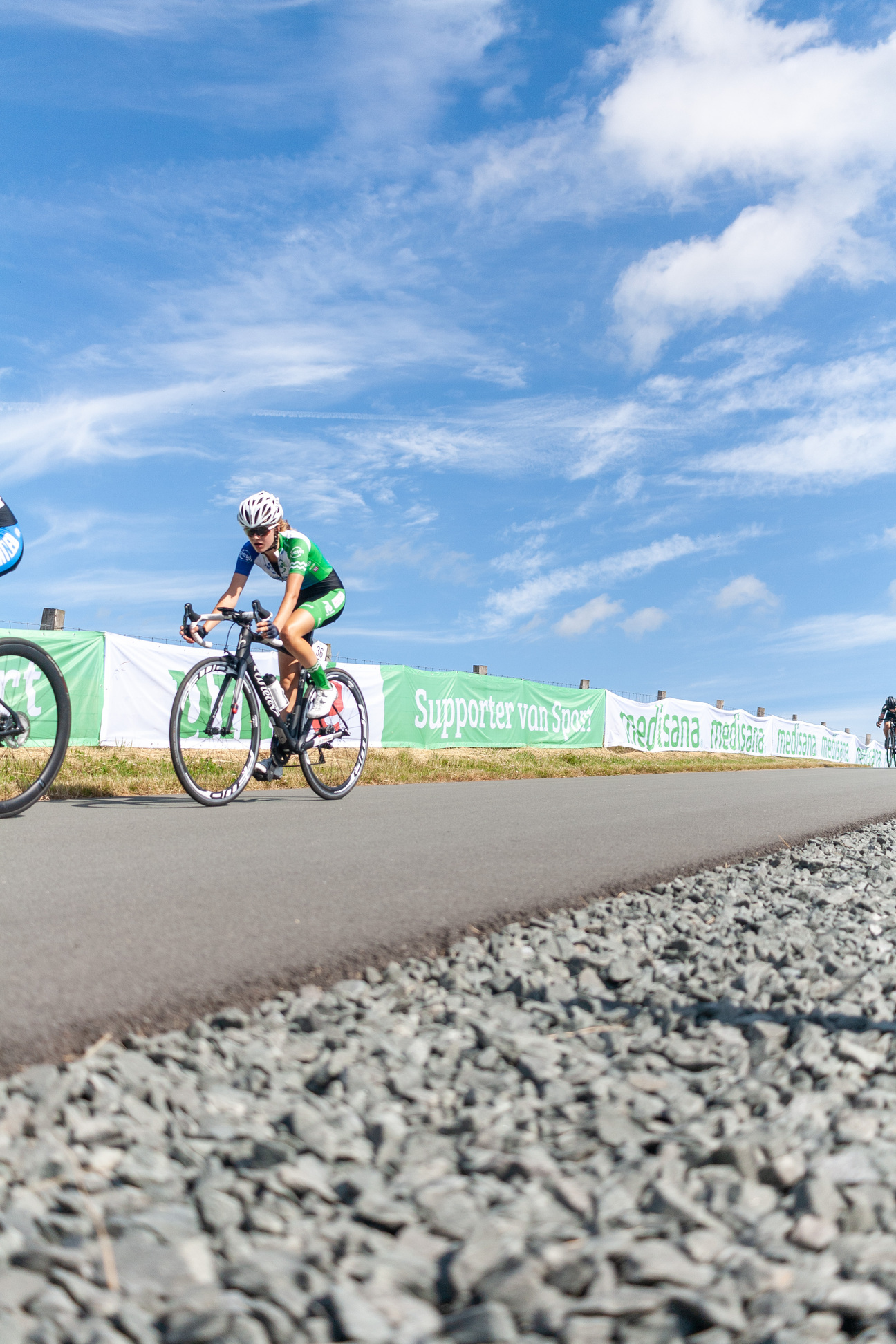 A cyclist in a green jersey and white helmet is riding on a gravel road at the Wielrennen Nieuwelingen Dames event.