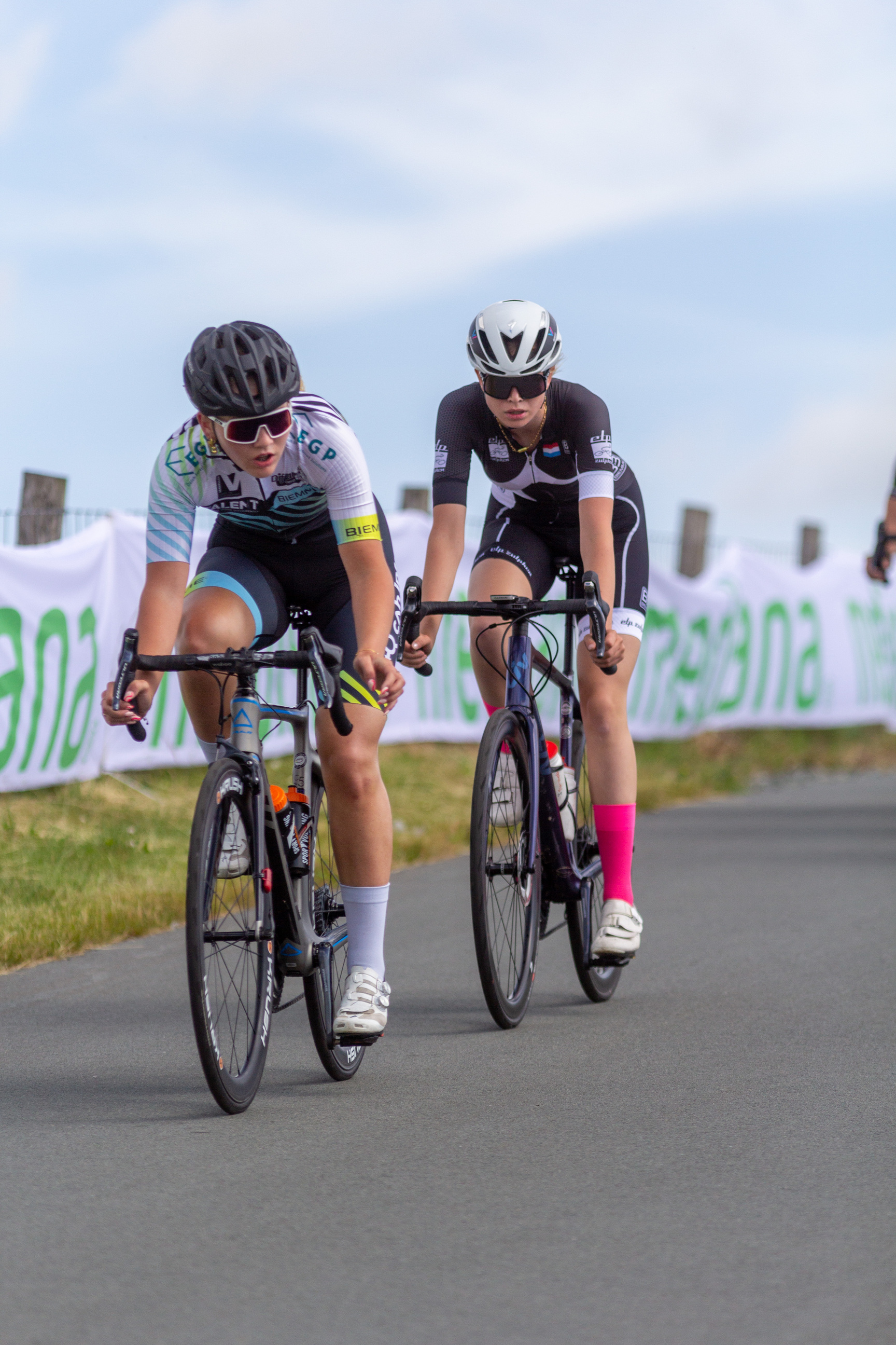 Two women riding bikes together on a road, one of which is wearing a white helmet.