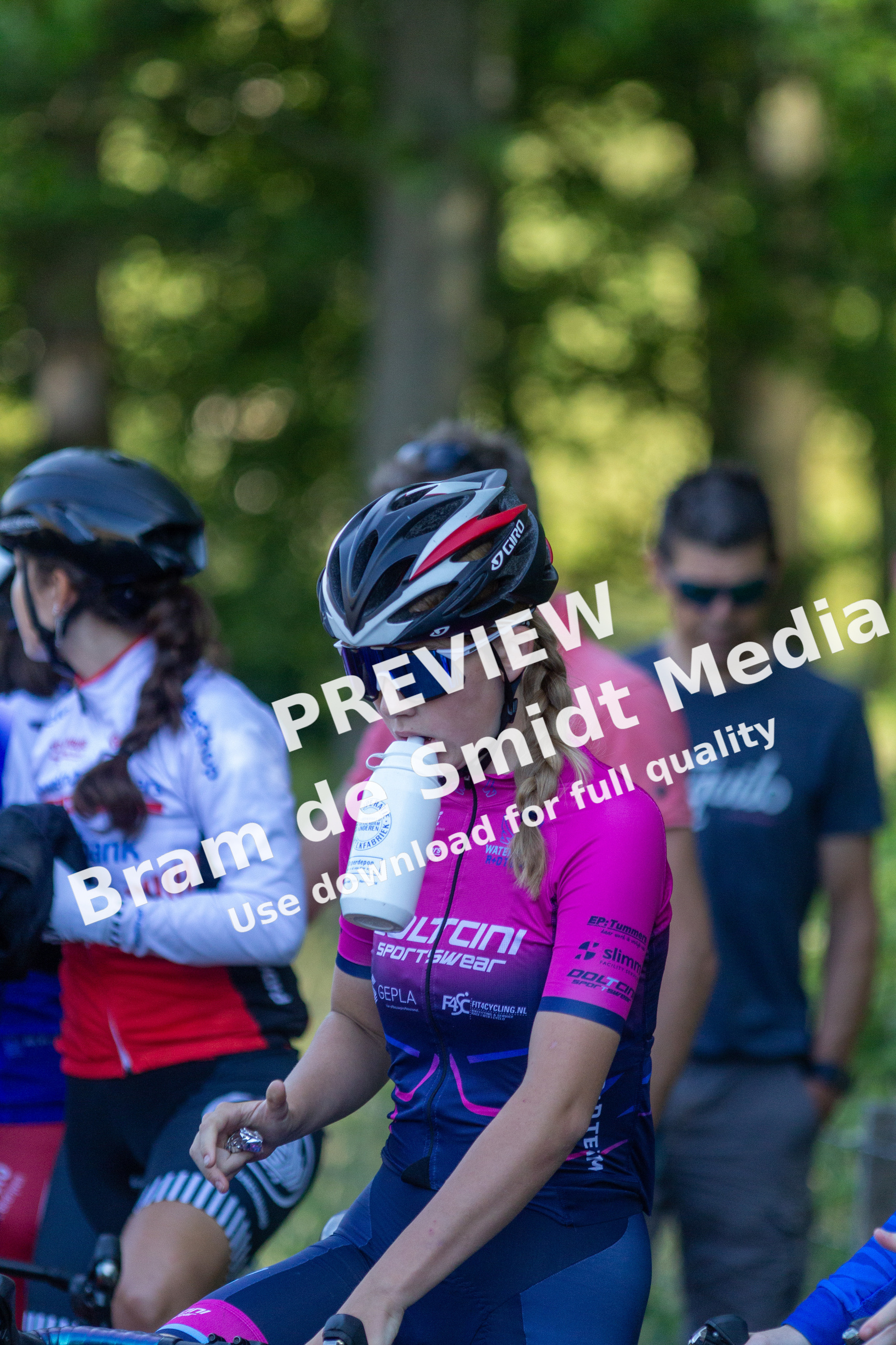 A young woman wearing a pink and blue cycling jersey is drinking from a white water bottle while sitting on a bicycle.