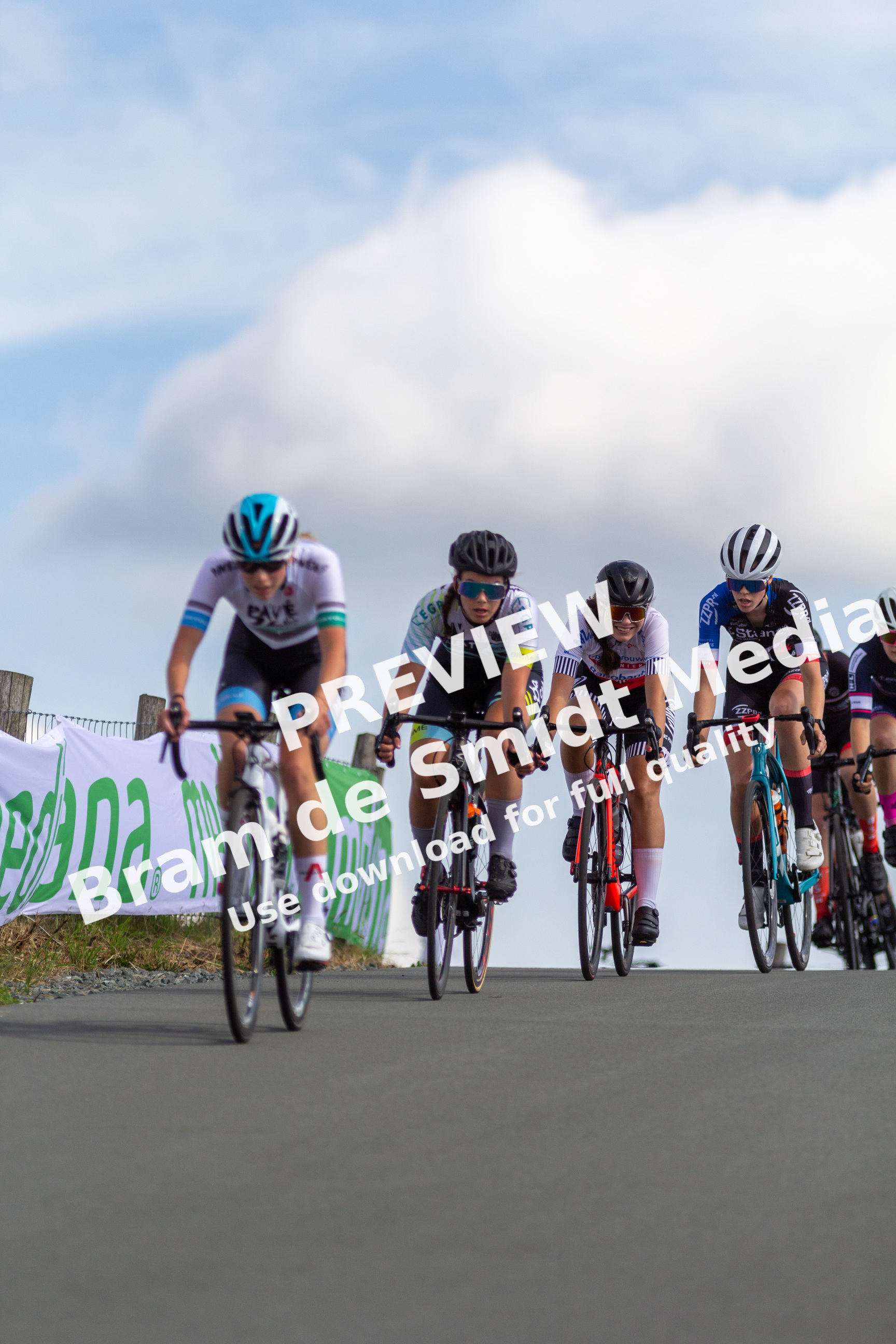 A group of bicyclists, sponsored by NK Dames, compete on a road during Wielrennen.