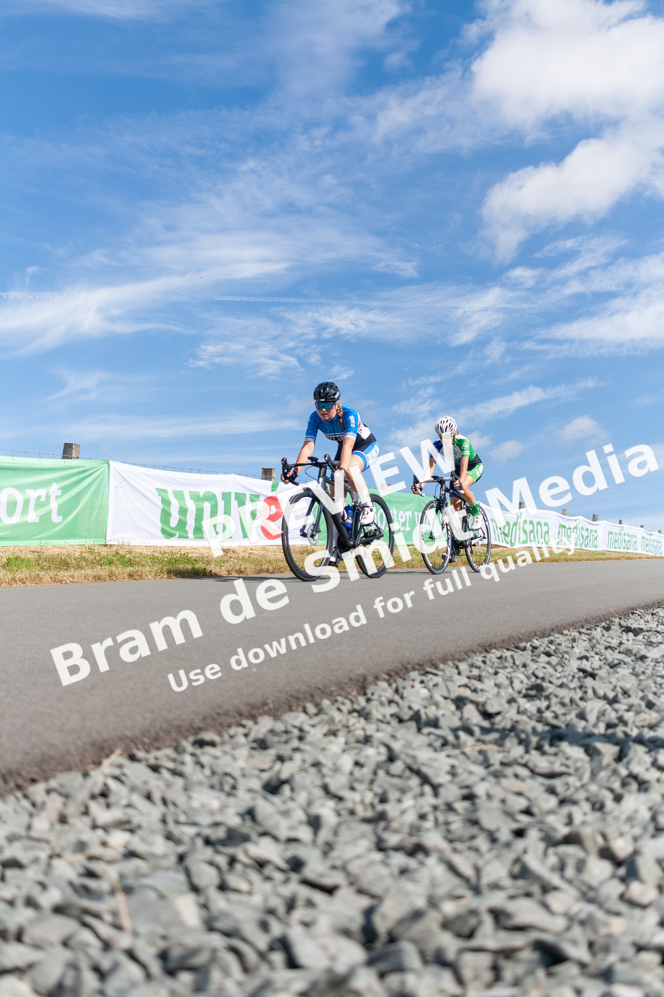 Two cyclists ride side by side on a gravel road with a banner displaying "University Sport" in the background.