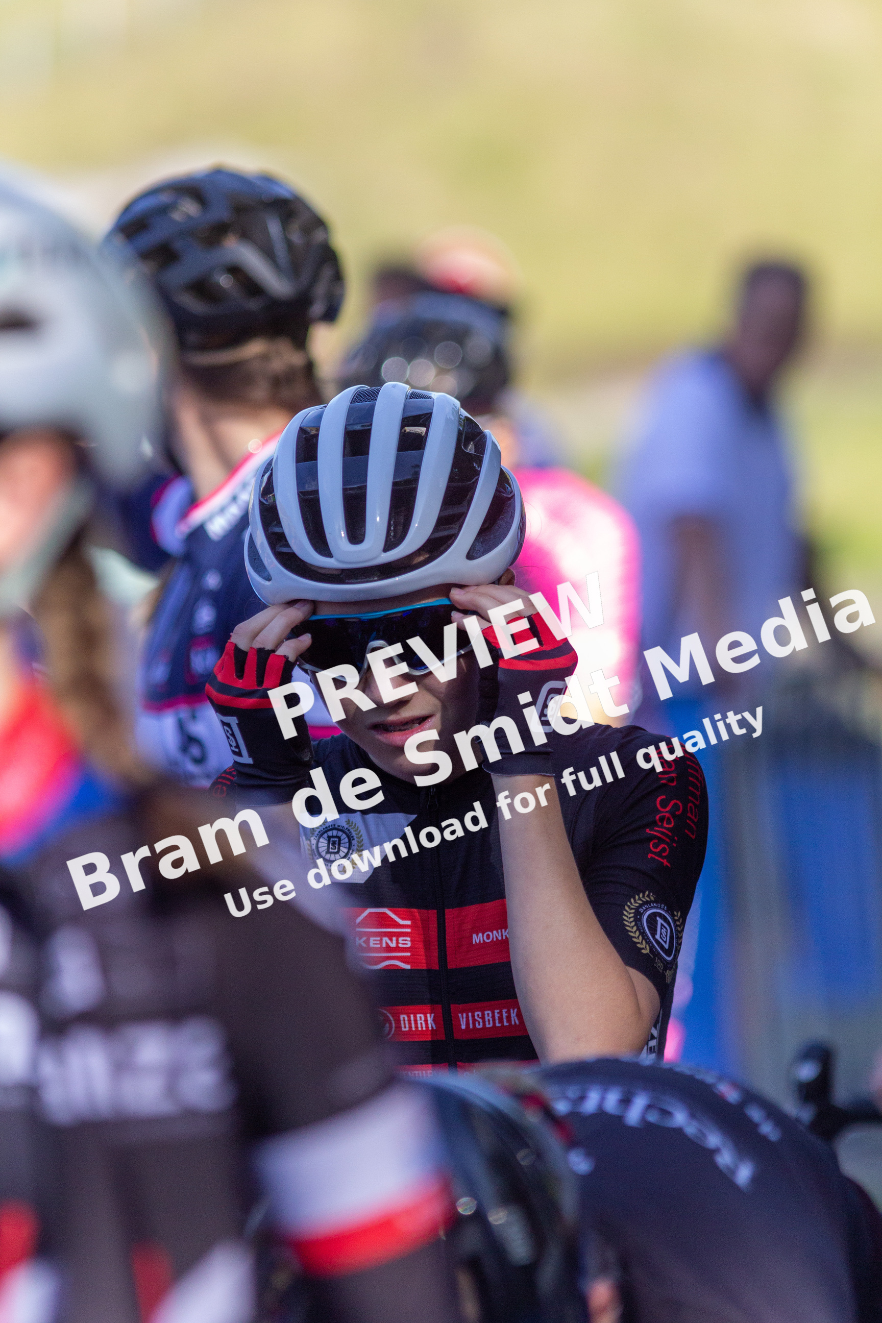 A group of cyclists with a red and black jersey on that says "NK Dames" are wearing helmets and sunglasses.