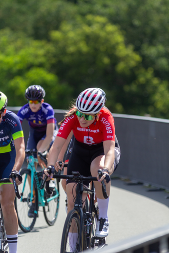 Two women on bikes with red and white shirts with the word weilrennen on them race each other.