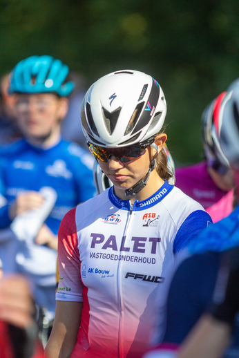 A lady wearing a white and red jersey is standing in front of two other cyclists. The jersey has the word "PALET" on it.