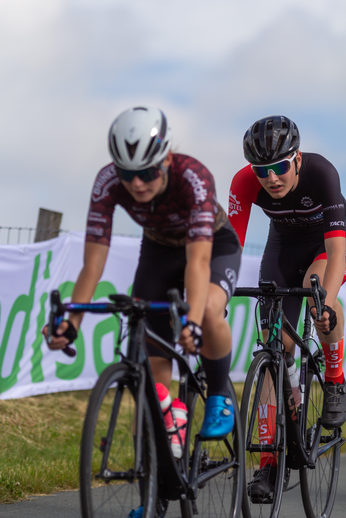 Two women in black and red cycling outfits are riding their bicycles on the road.