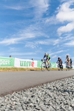 A group of cyclists race on a paved road with an ad that says "Sport" in the background.