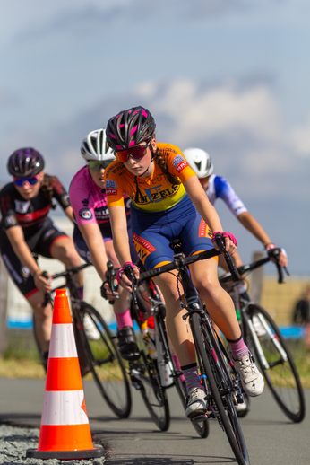 Four women wearing helmets, one of them with a pink helmet, race on their bikes.