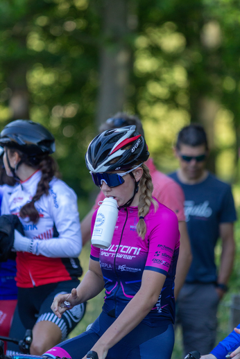 A young woman wearing a pink and blue cycling jersey is drinking from a white water bottle while sitting on a bicycle.