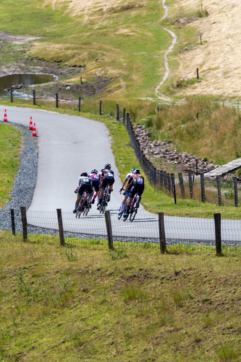 A group of cyclists are riding down a road that's next to a fence.