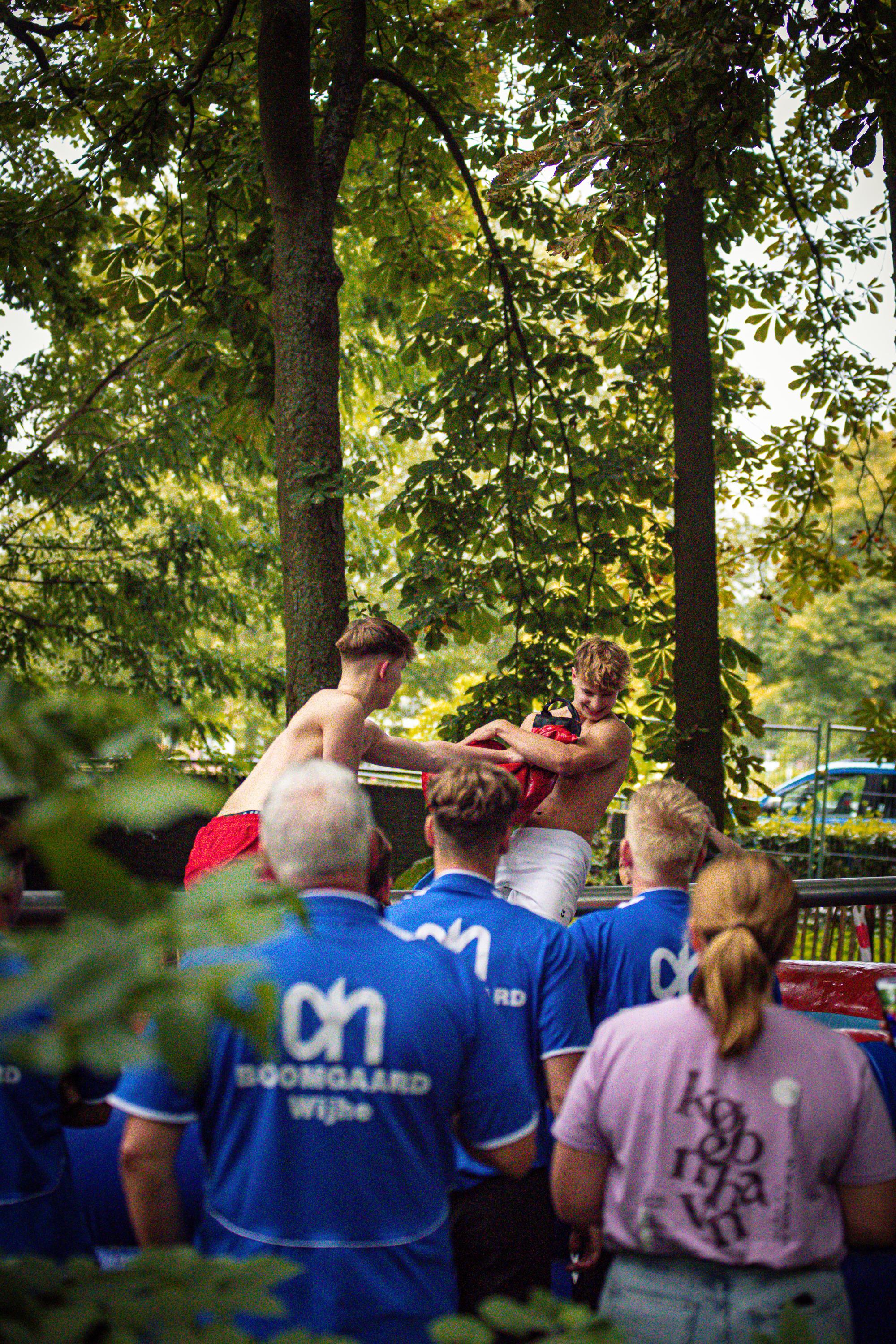 A boy wrestles a man in front of an audience in blue shirts that say "Zekerheid".