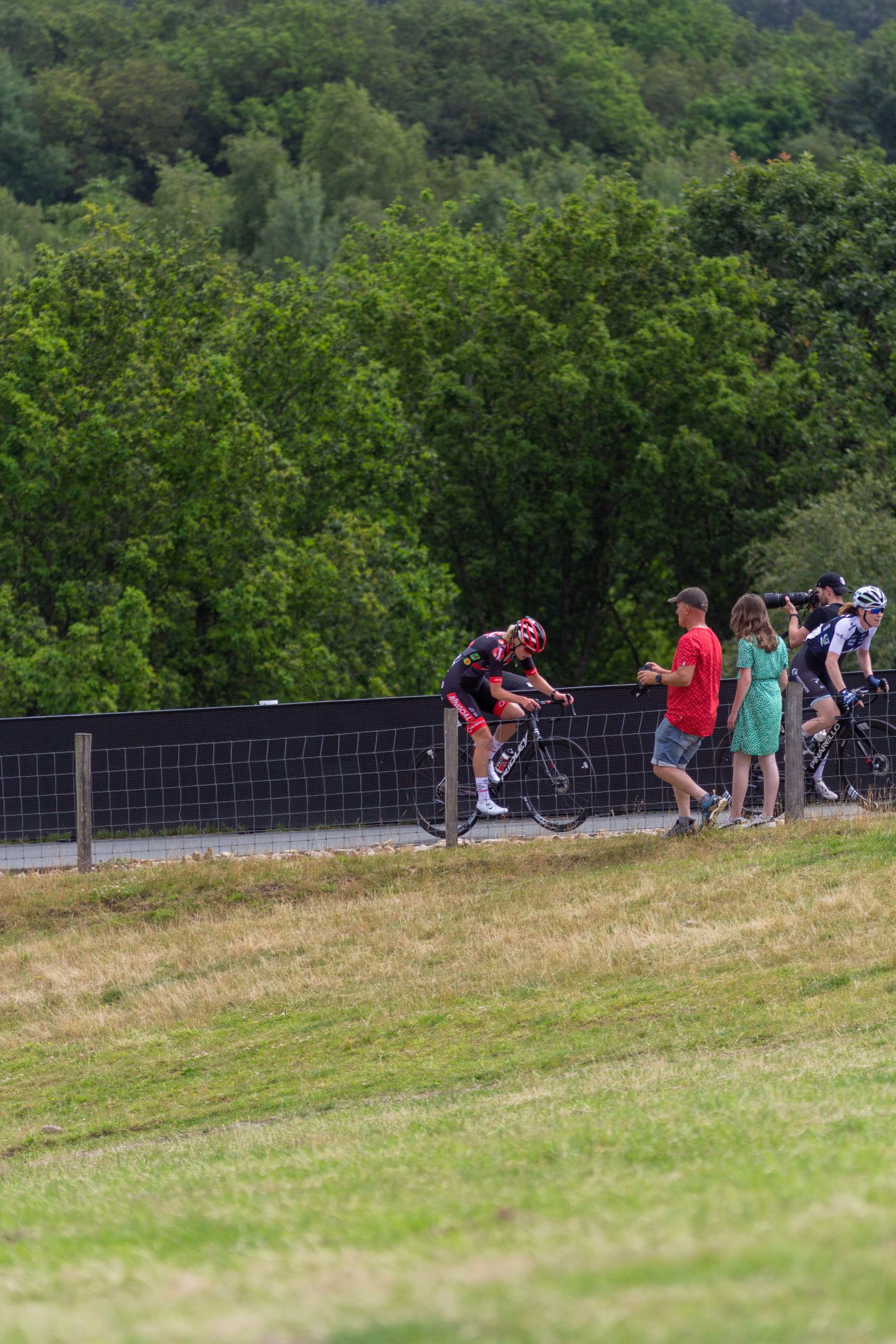 A couple of people are standing next to each other, watching two cyclists ride on a road.