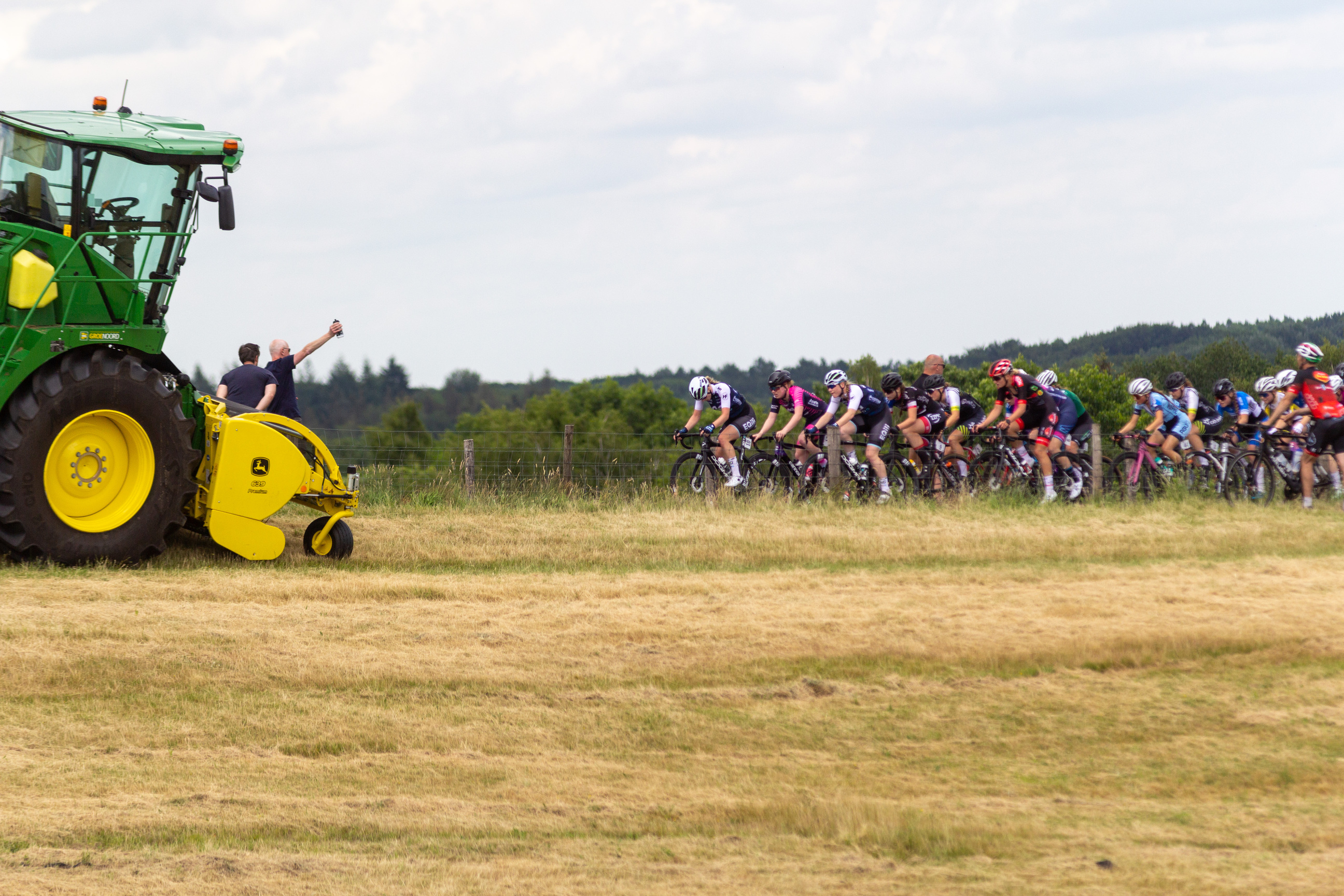 A man holding up his arm next to a tractor while a group of cyclists ride through the field.