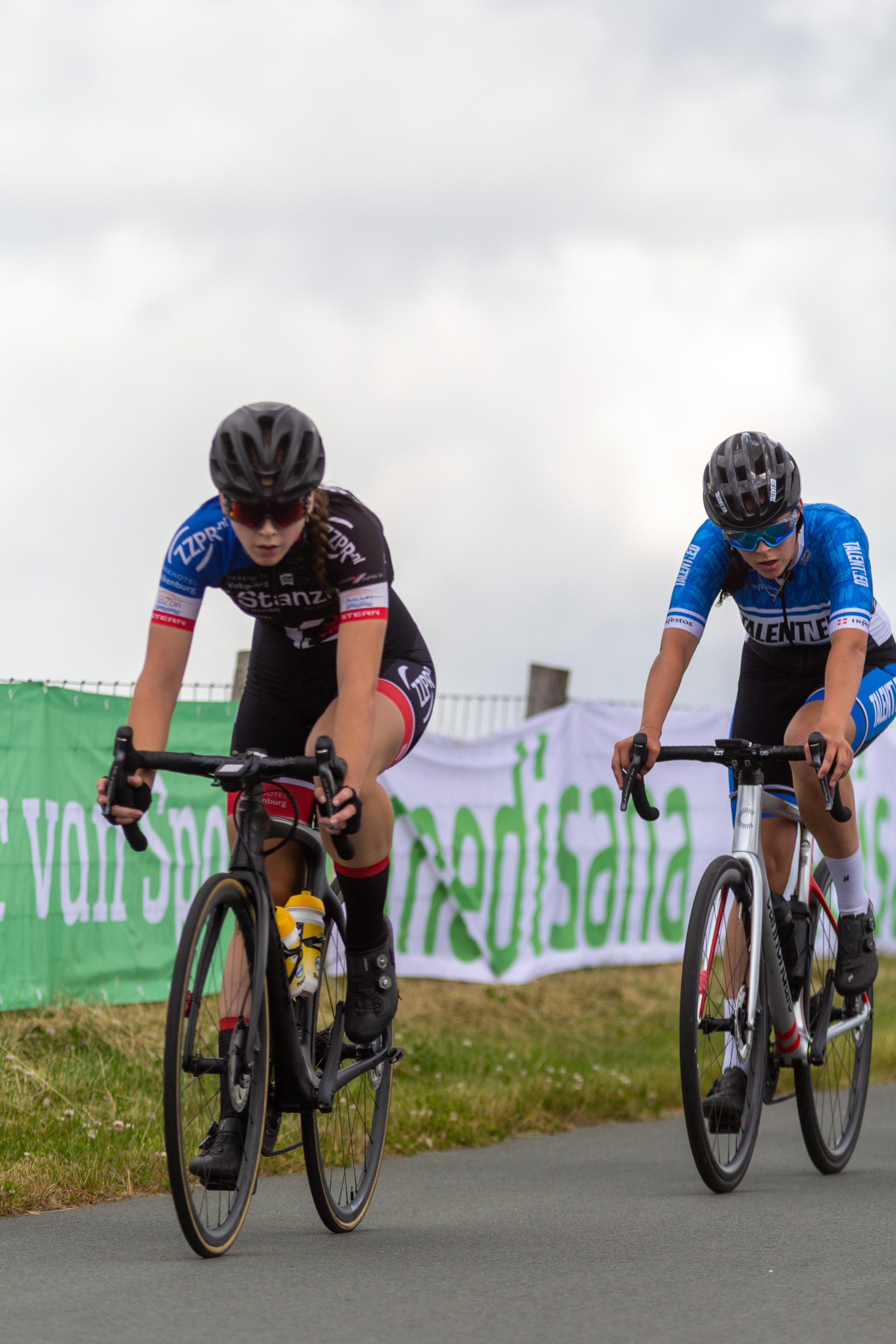 "Two people ride bicycles on a road with a banner that reads Mediaset in the background".