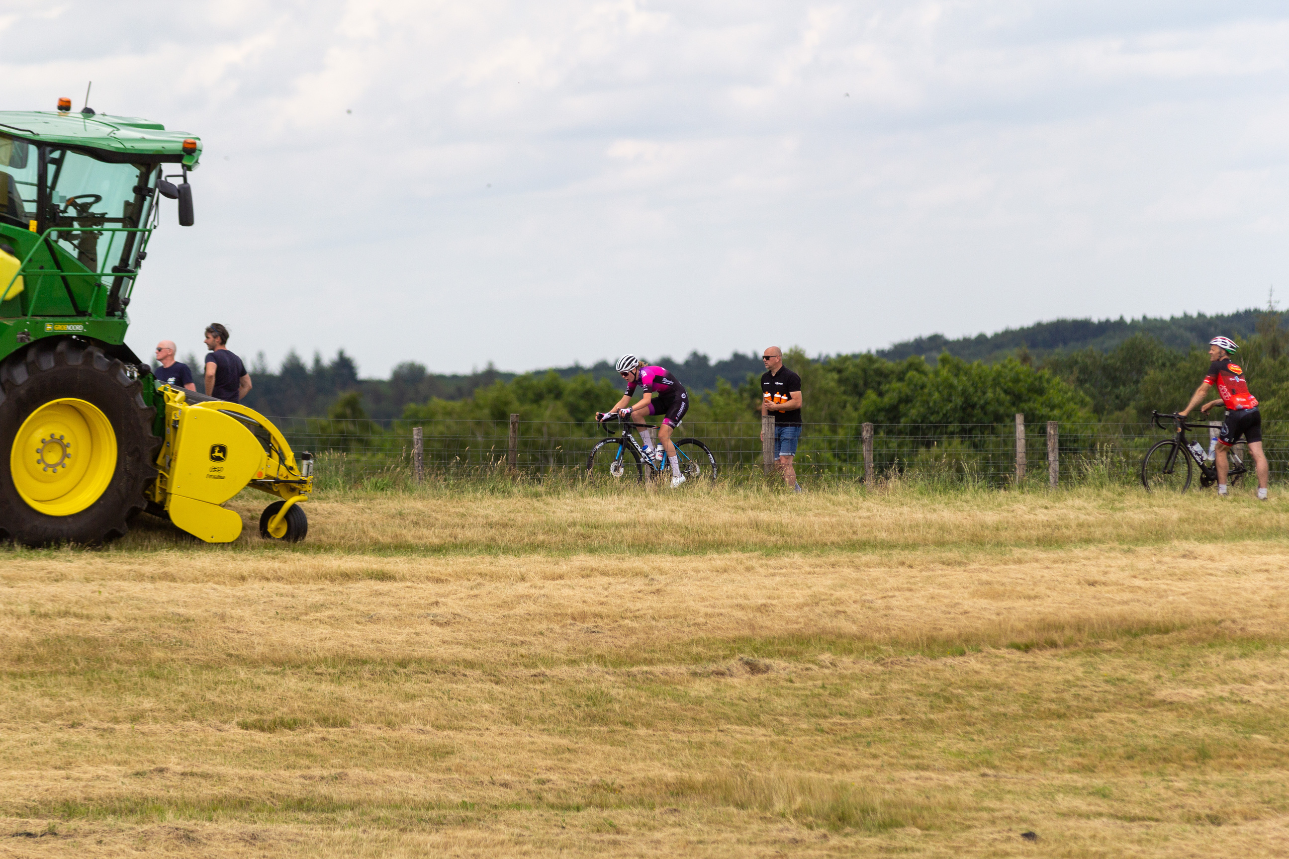 Junioren Dames ride bikes in a field as an orange tractor pulls up.