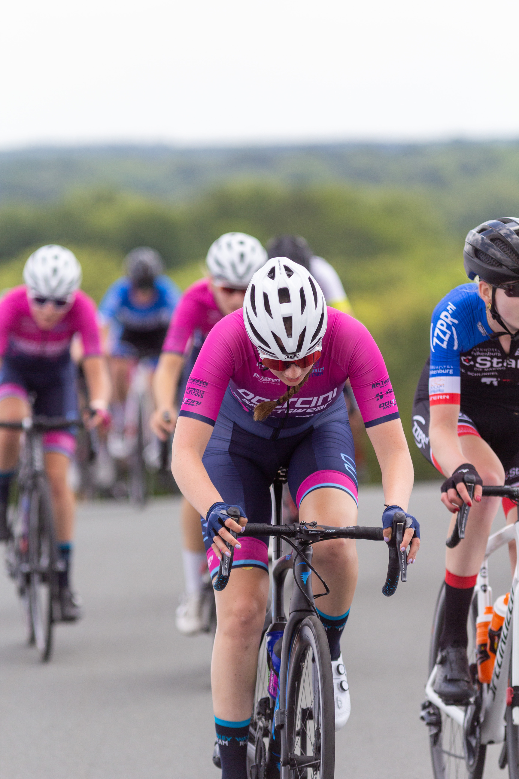 A group of female cyclists are wearing blue, pink and black jerseys as they ride their bikes on the road.