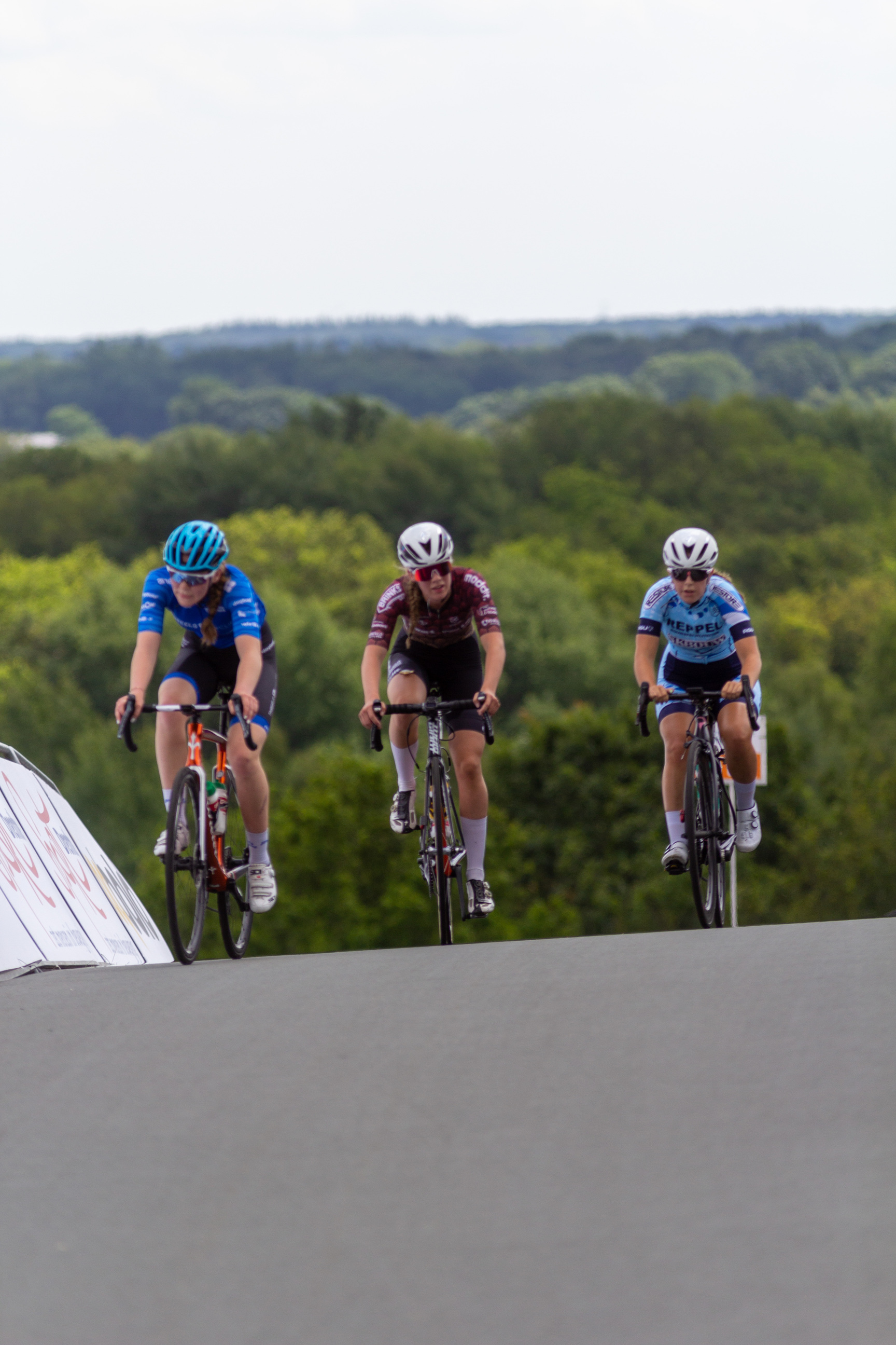 Three women race on bikes wearing helmets and the lead woman has a blue helmet.