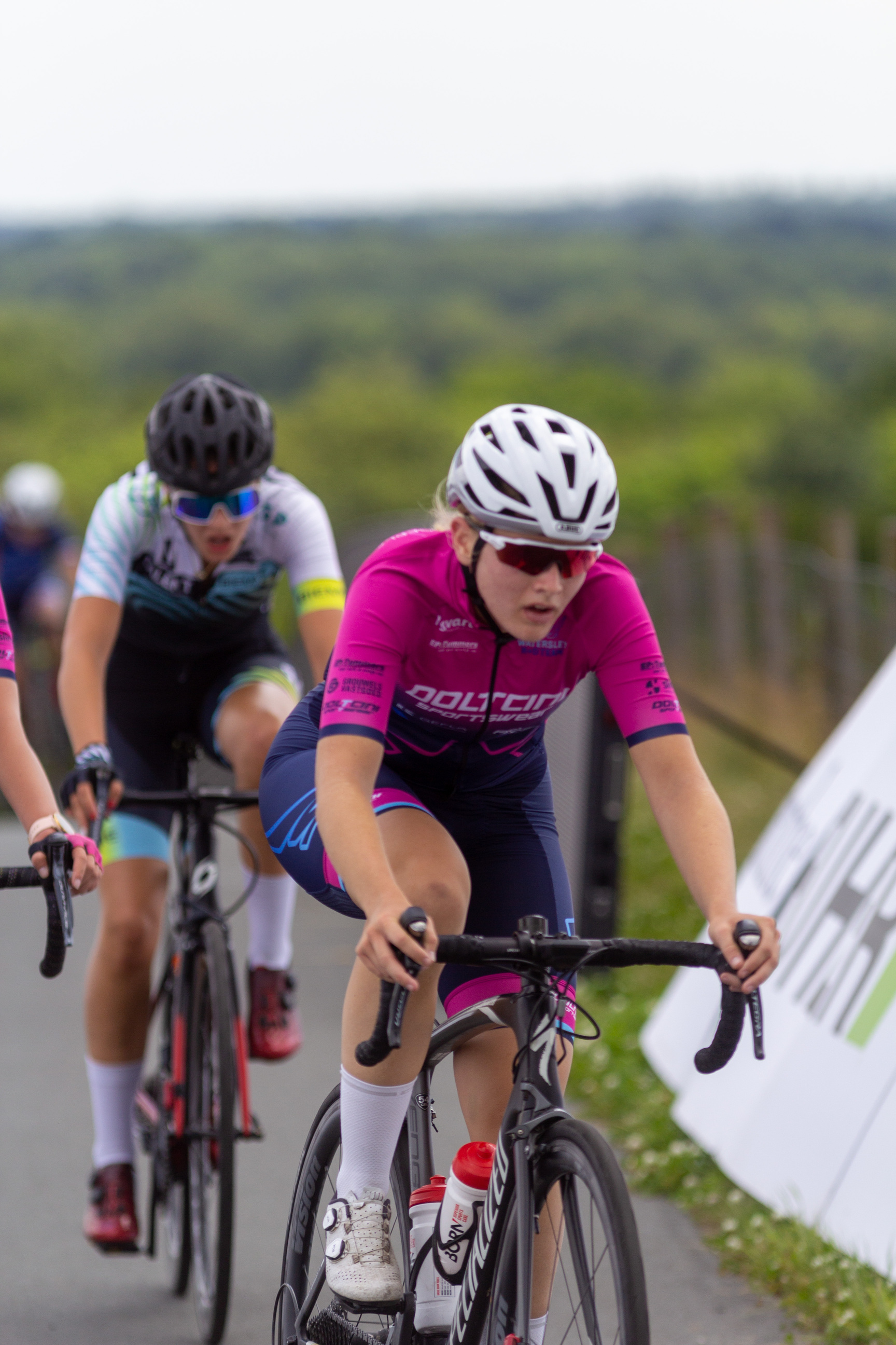 Two female cyclists in a race are wearing black helmets and pink jerseys.