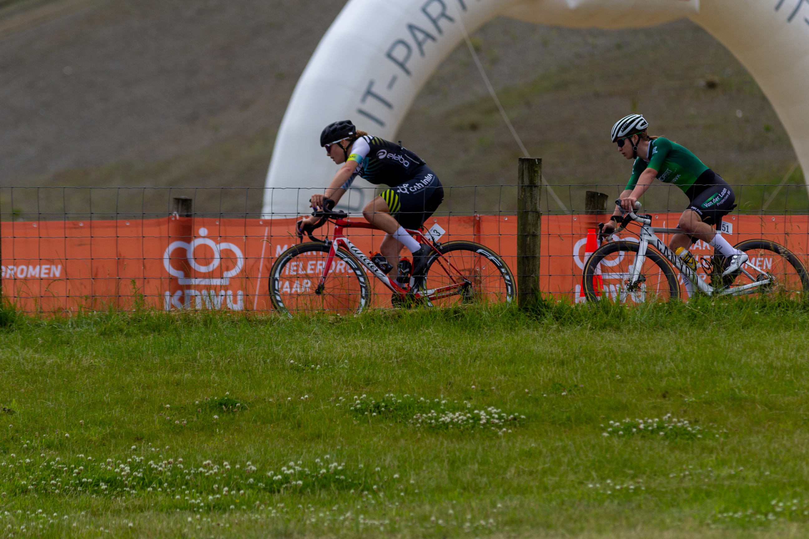 Two men riding on bikes in front of an orange sign that says "Junioren Dames".