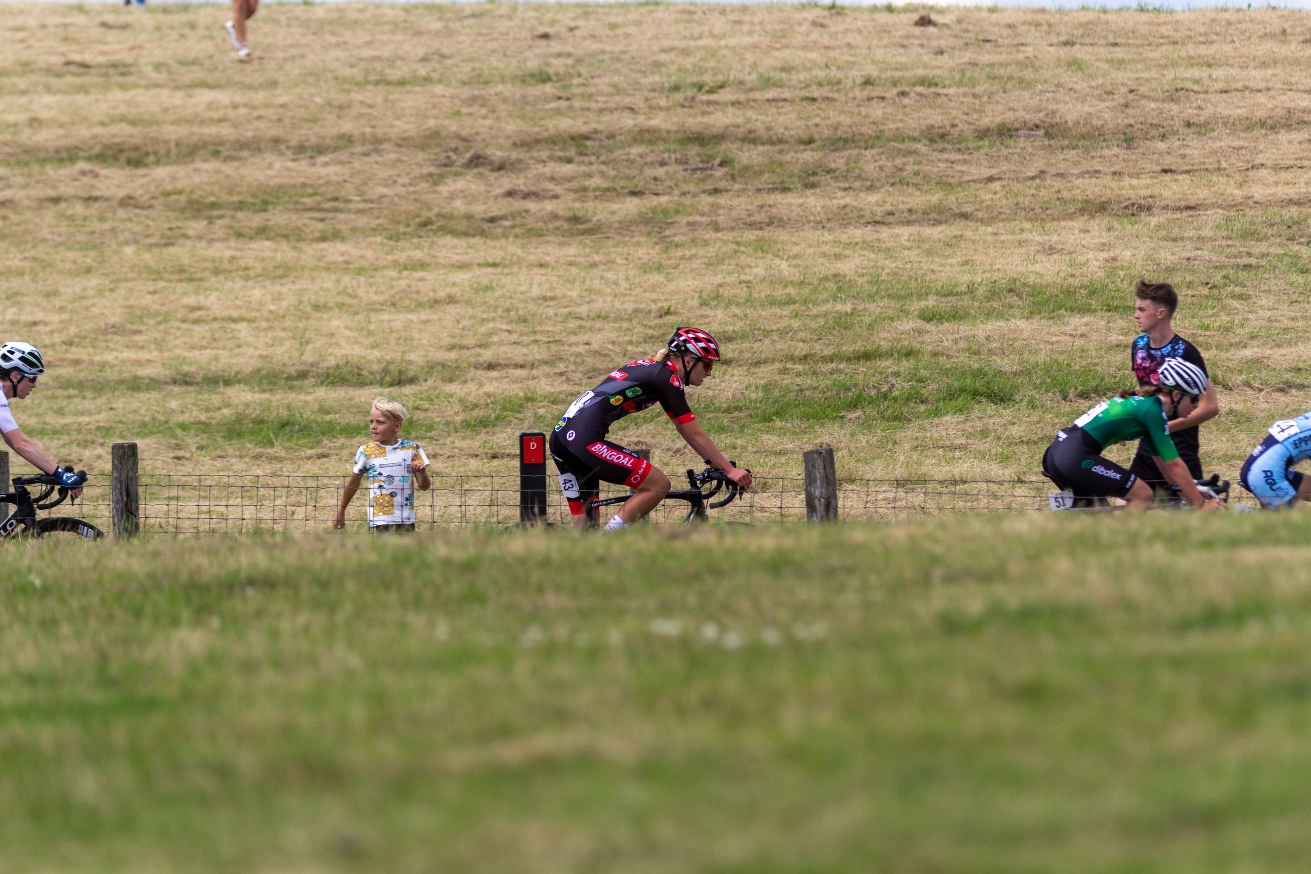 Two men riding bikes on a grassy field while one man and child look on.