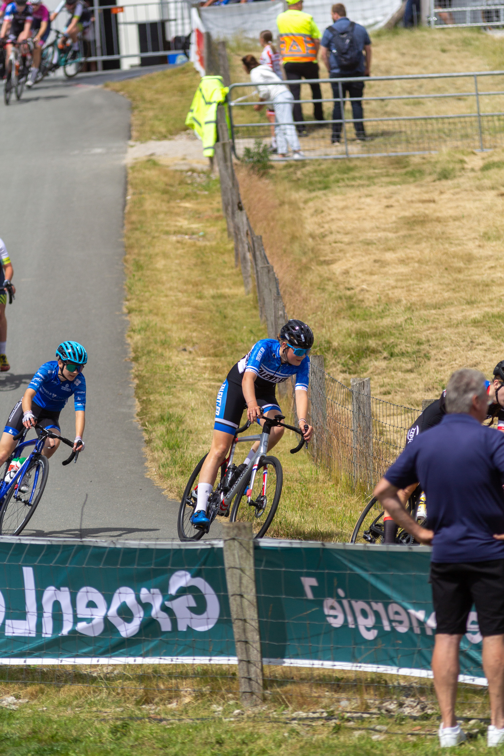 A cyclist in a blue and white uniform wearing a helmet is riding down a dirt road during the NK Dames Wielrennen.