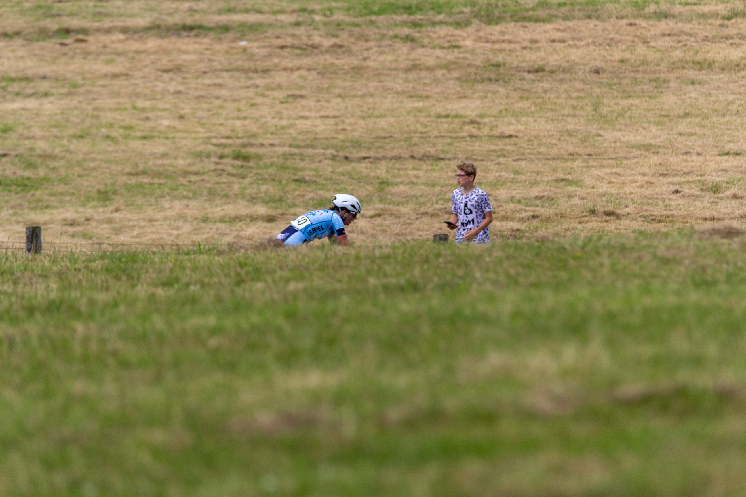 A young boy in a blue shirt and helmet sits on the grass as he watches a race.