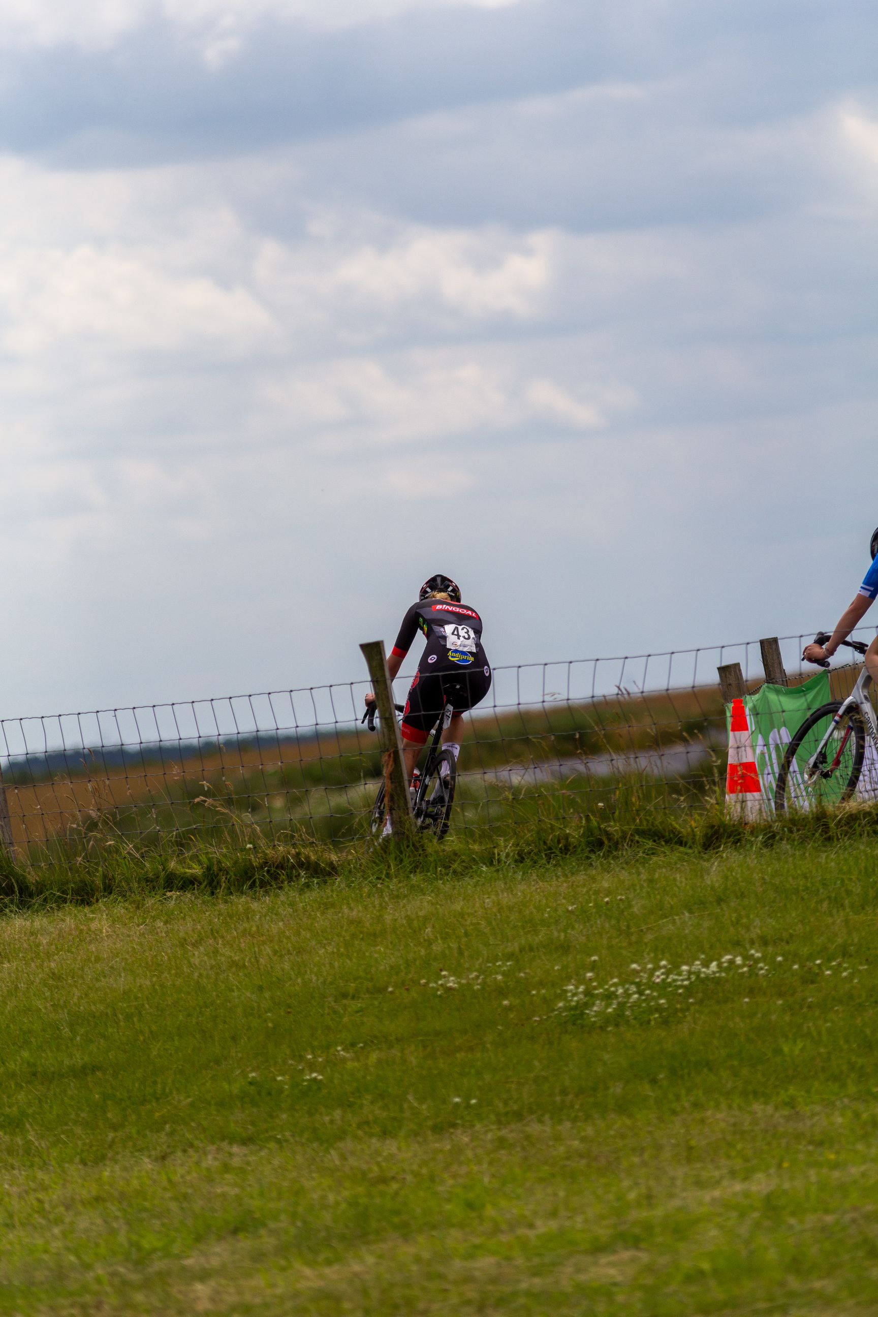 A cyclist in a red and black uniform is riding her bike near a fence. The fence has signs that say "Junioren Dames".