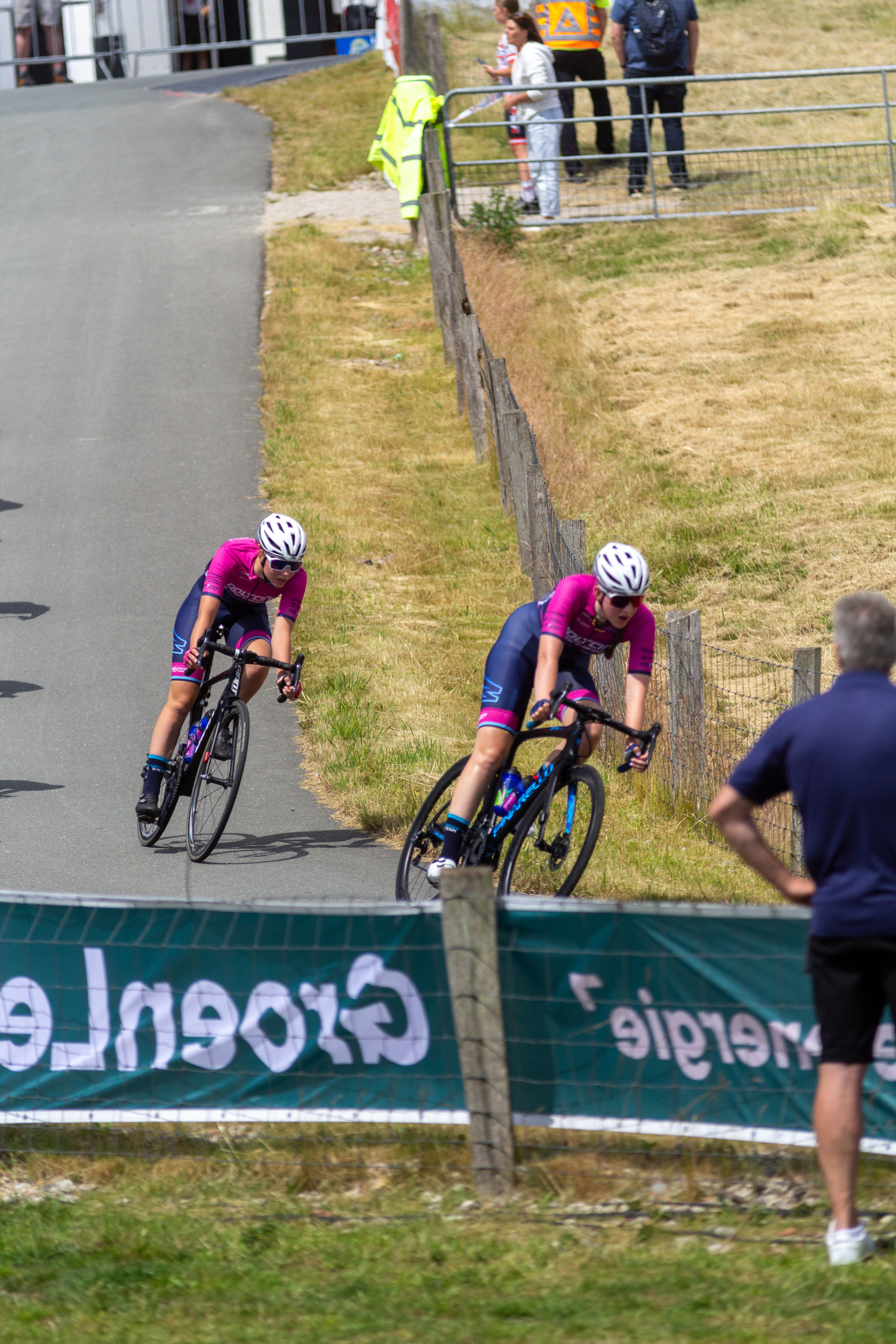 Two women are racing bicycles on a track, with one wearing a pink shirt and blue pants.