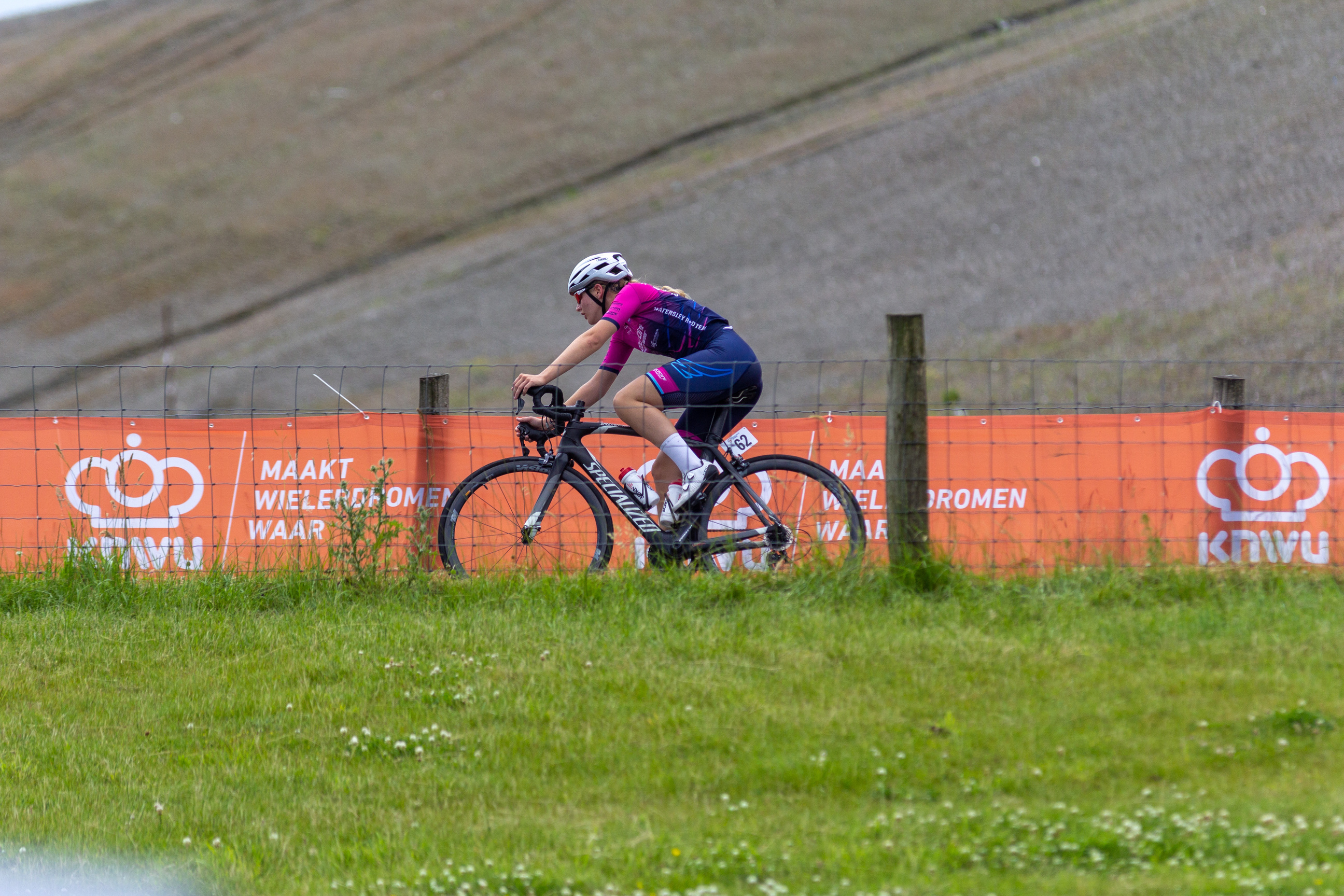 A man on a bike with a blue helmet and pink jersey in front of a fence that says KONVU.