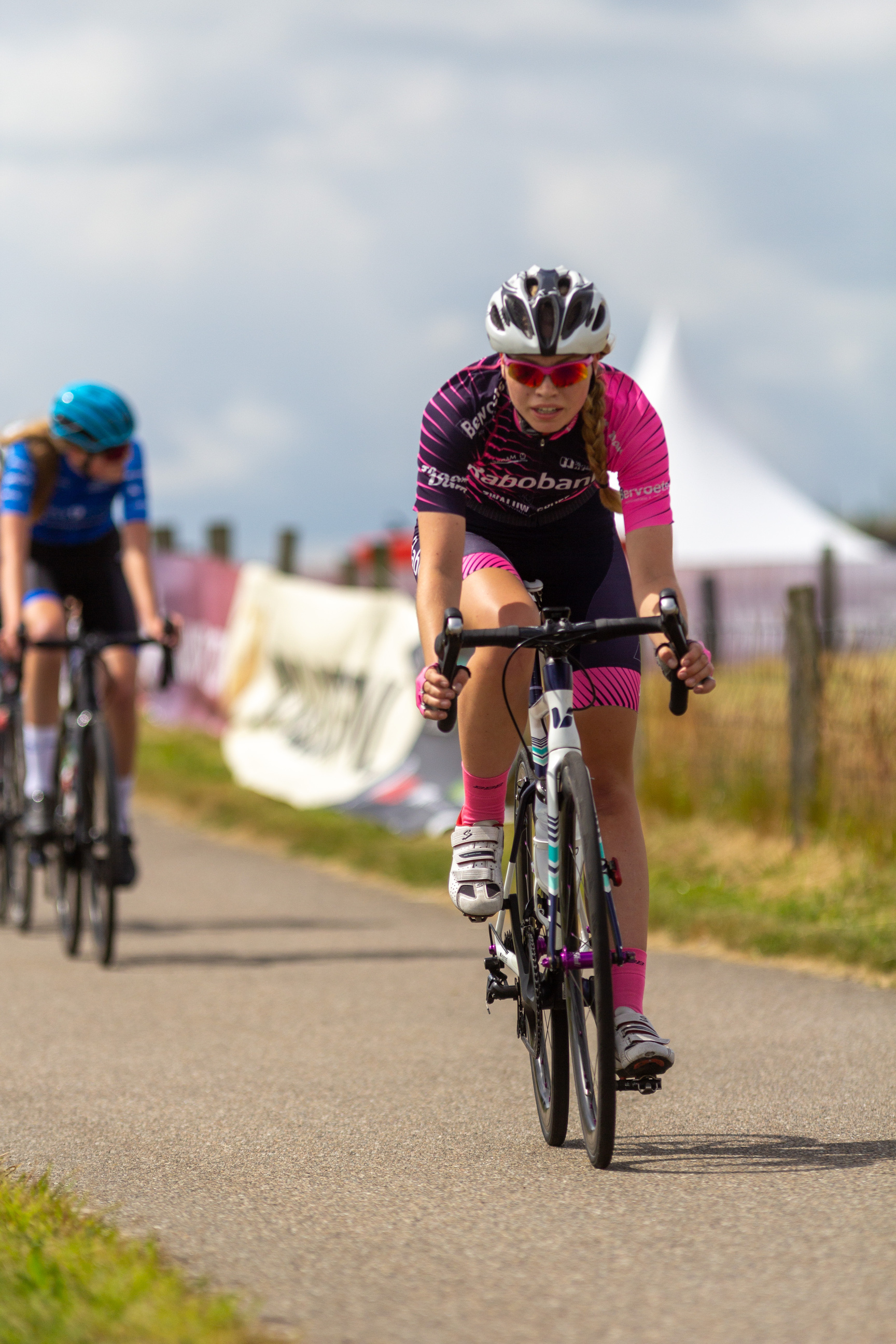 Two cyclists, a woman in pink and black with the number 2022 on her jersey is riding down a road.