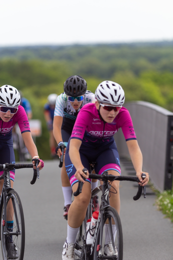 Three women wearing helmets and cycling jerseys are on their bikes.