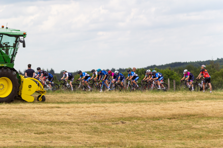 A green tractor pulls a yellow machine while children on bikes follow.