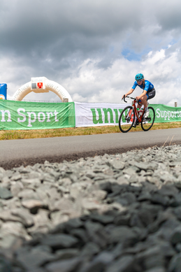 A cyclist in a blue shirt is riding on gravel near a green banner that says Wielrennen.