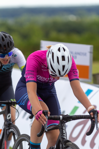 A woman wearing a purple shirt and helmet is riding a bike on the track with another person behind her.