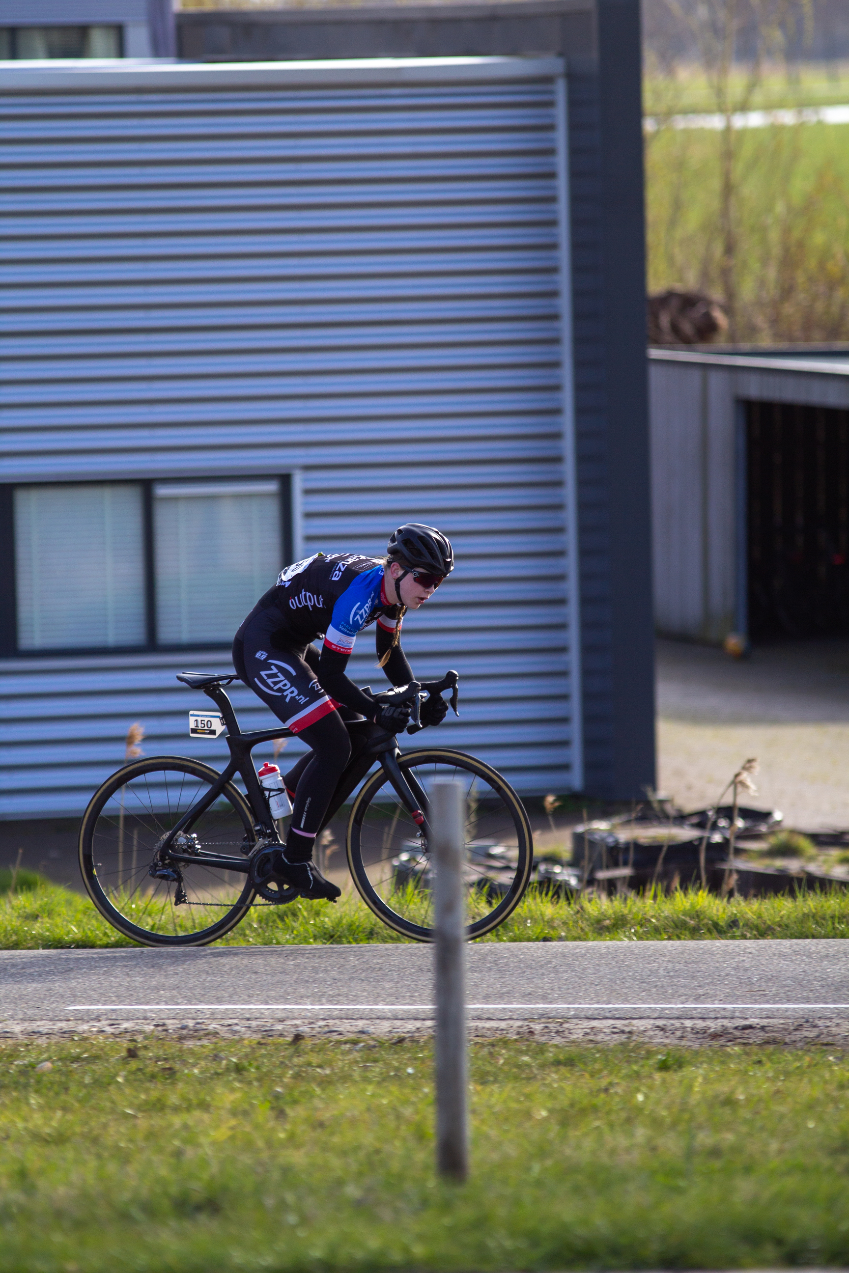 A cyclist wearing a blue and white top and black pants rides down the road, passing by a small shop.