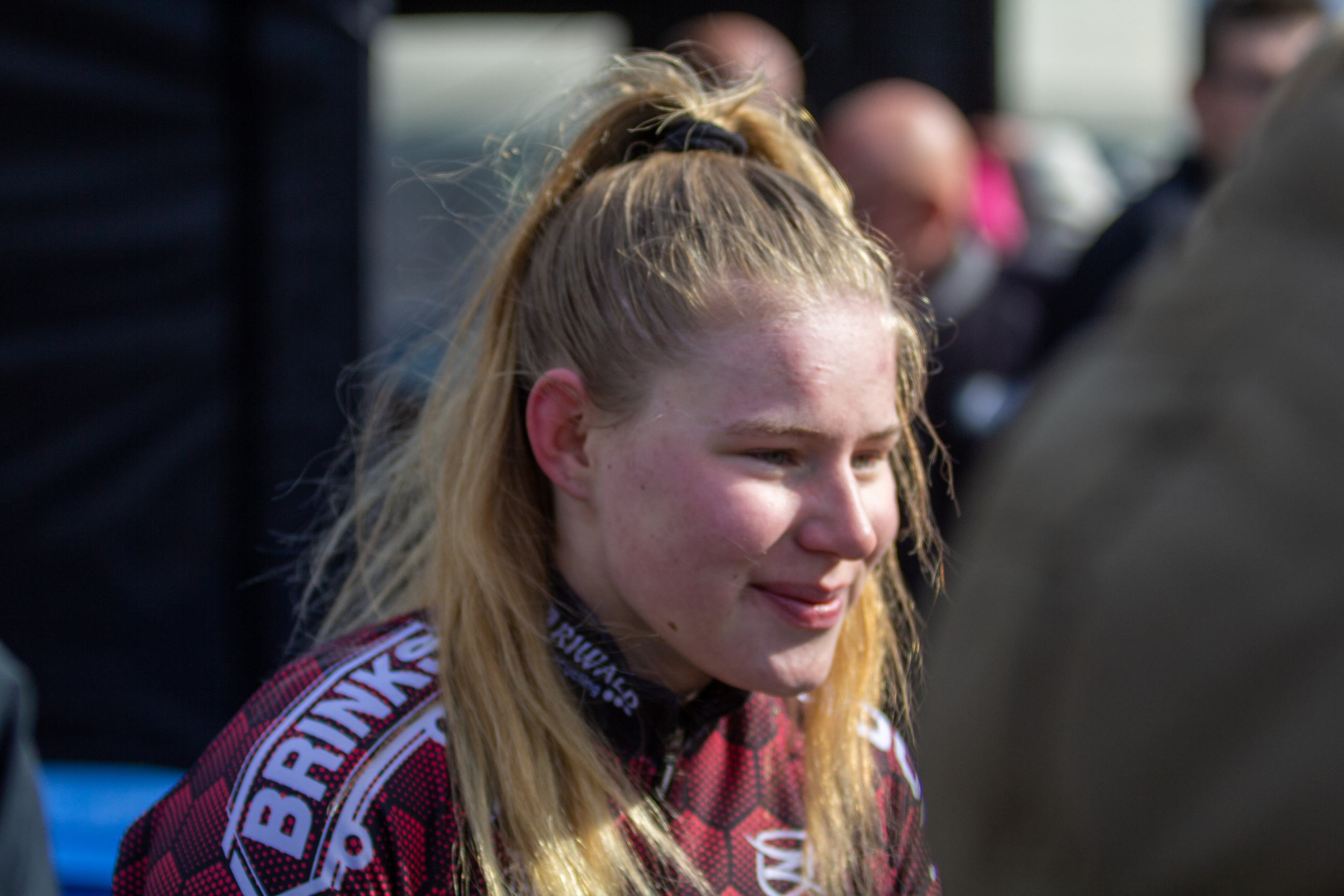 A woman with blonde hair and a white shirt is wearing a red and black cycling jersey. The jersey has the word "Brinkz" on it.