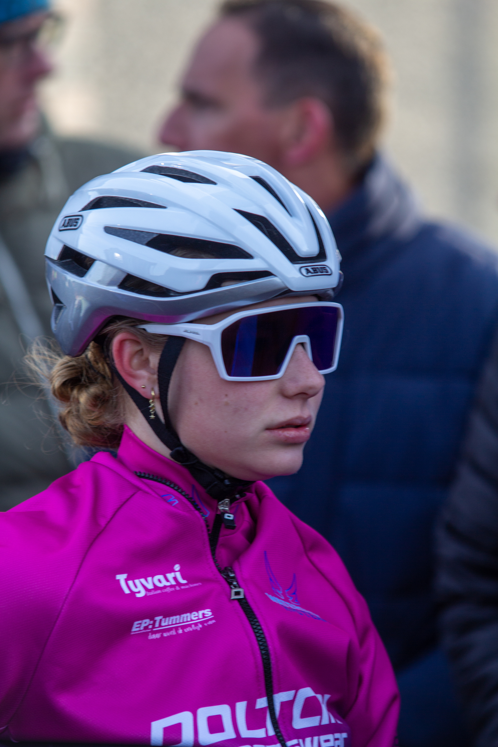 A young woman in a purple jersey is wearing a white bike helmet with black and blue details. She has blonde hair.