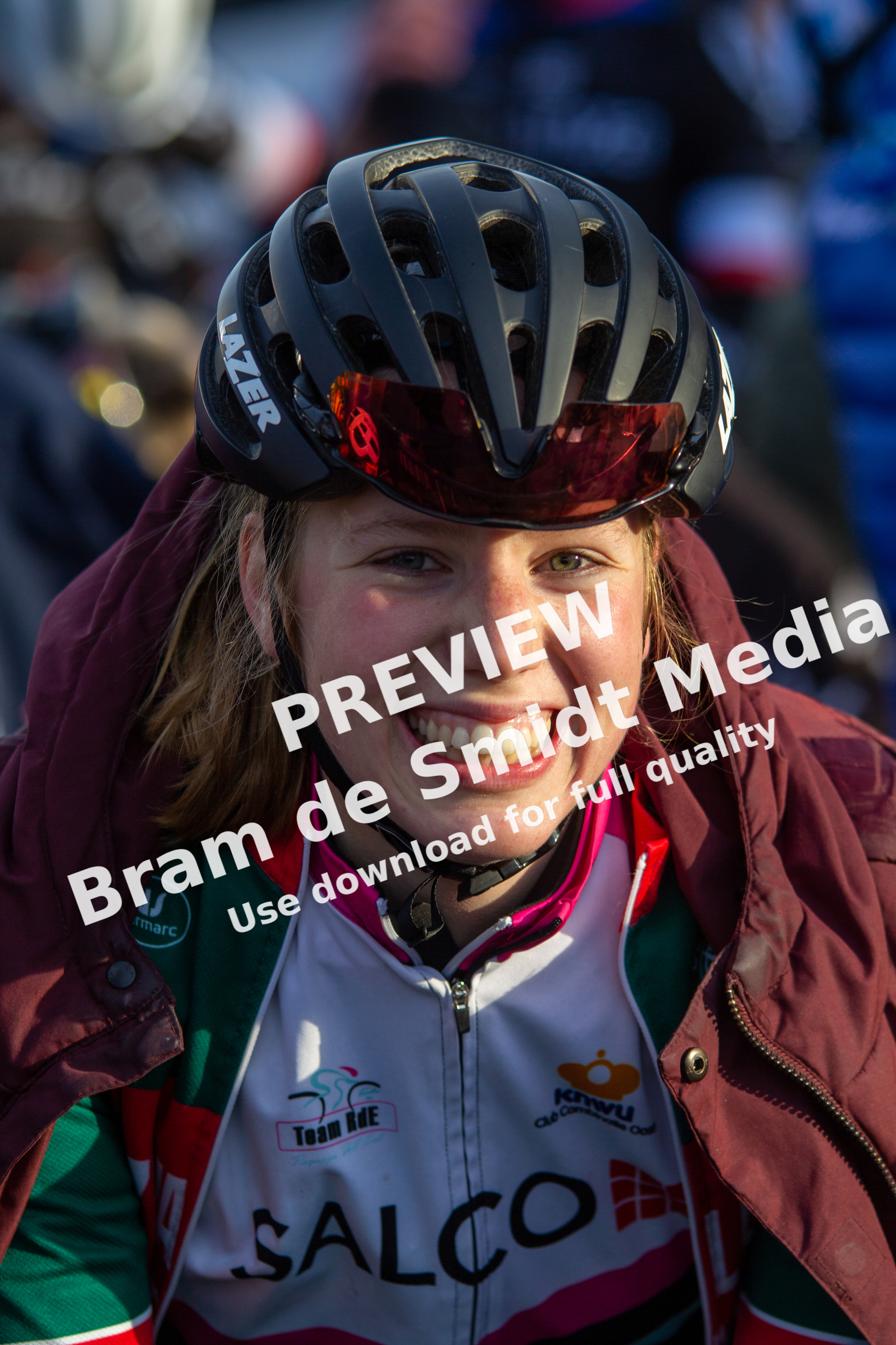 A girl is wearing a helmet and smiling, with a bike race in the background. She has a red jacket and her shirt says "SALCO".