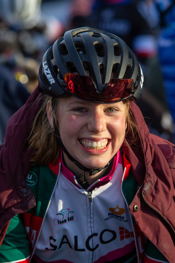 A girl is wearing a helmet and smiling, with a bike race in the background. She has a red jacket and her shirt says "SALCO".