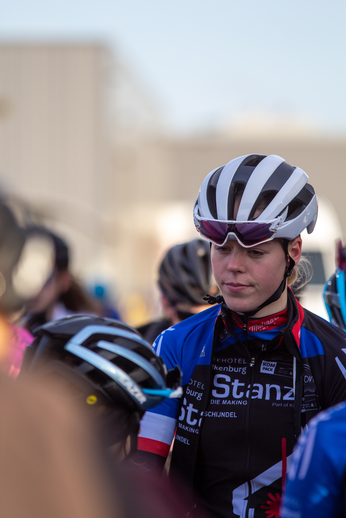 A cyclist wearing a blue jersey with "St. Anne" on the front is surrounded by other cyclists in the background.