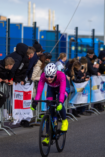 A cyclist wearing a pink outfit is riding down the street with spectators watching from behind a metal barricade.