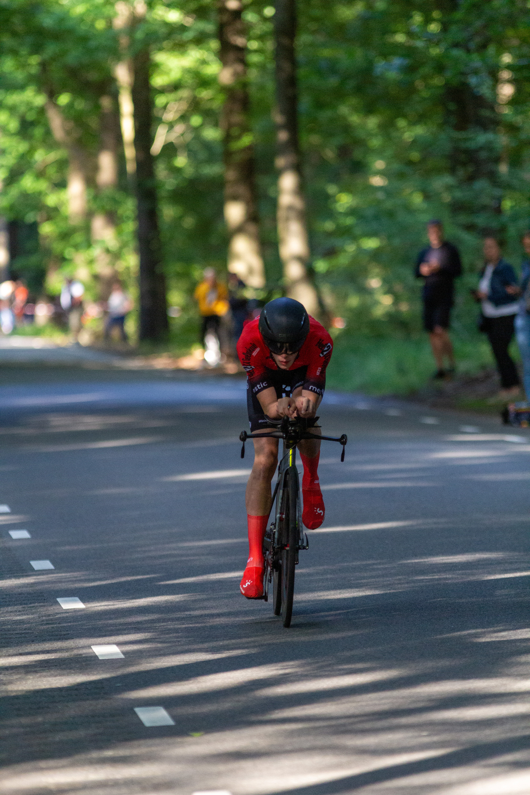 A cyclist races on a road lined with trees, wearing red shoes and a helmet.