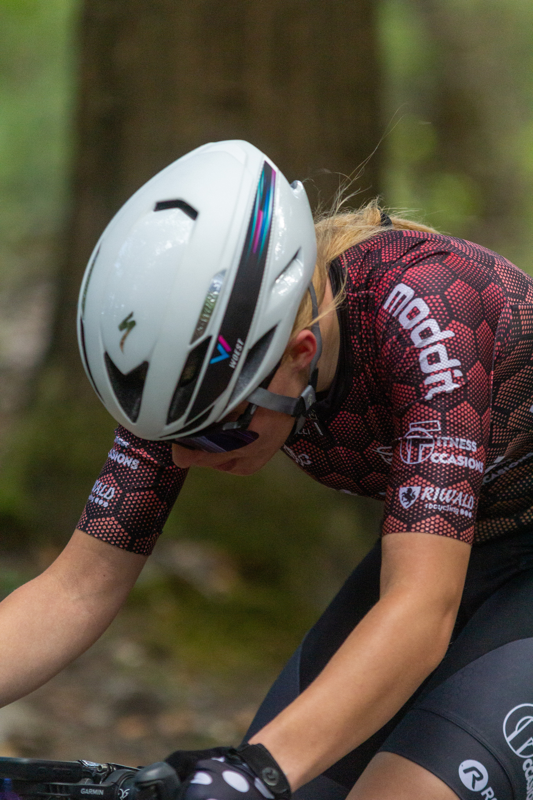 A woman is wearing a black and red cycling jersey while riding her bike outdoors. She also has a white helmet on for safety.