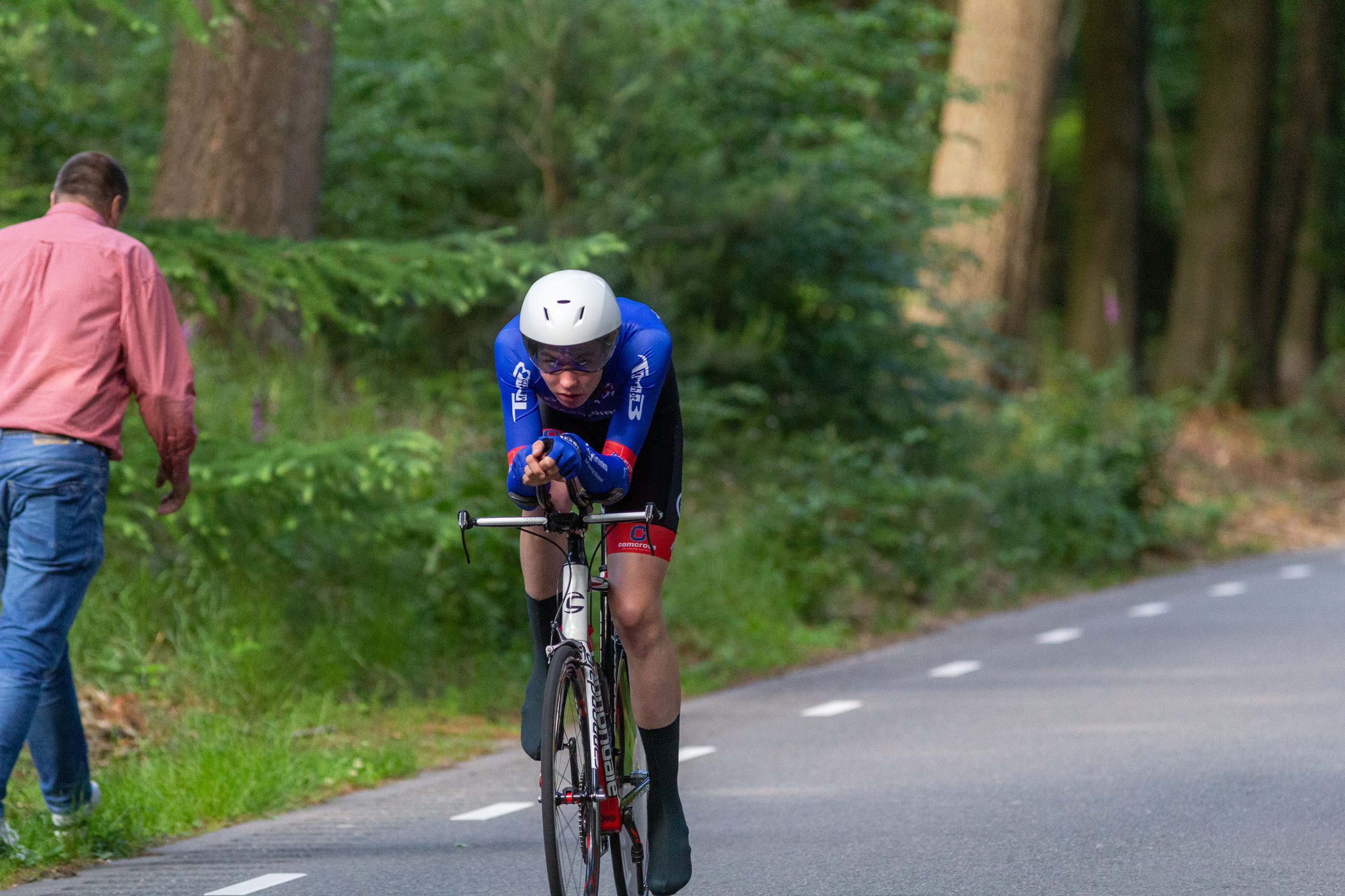 A cyclist wearing a blue and black jersey is on a road, surrounded by trees. The cyclist's helmet has the letters "DE" on it.