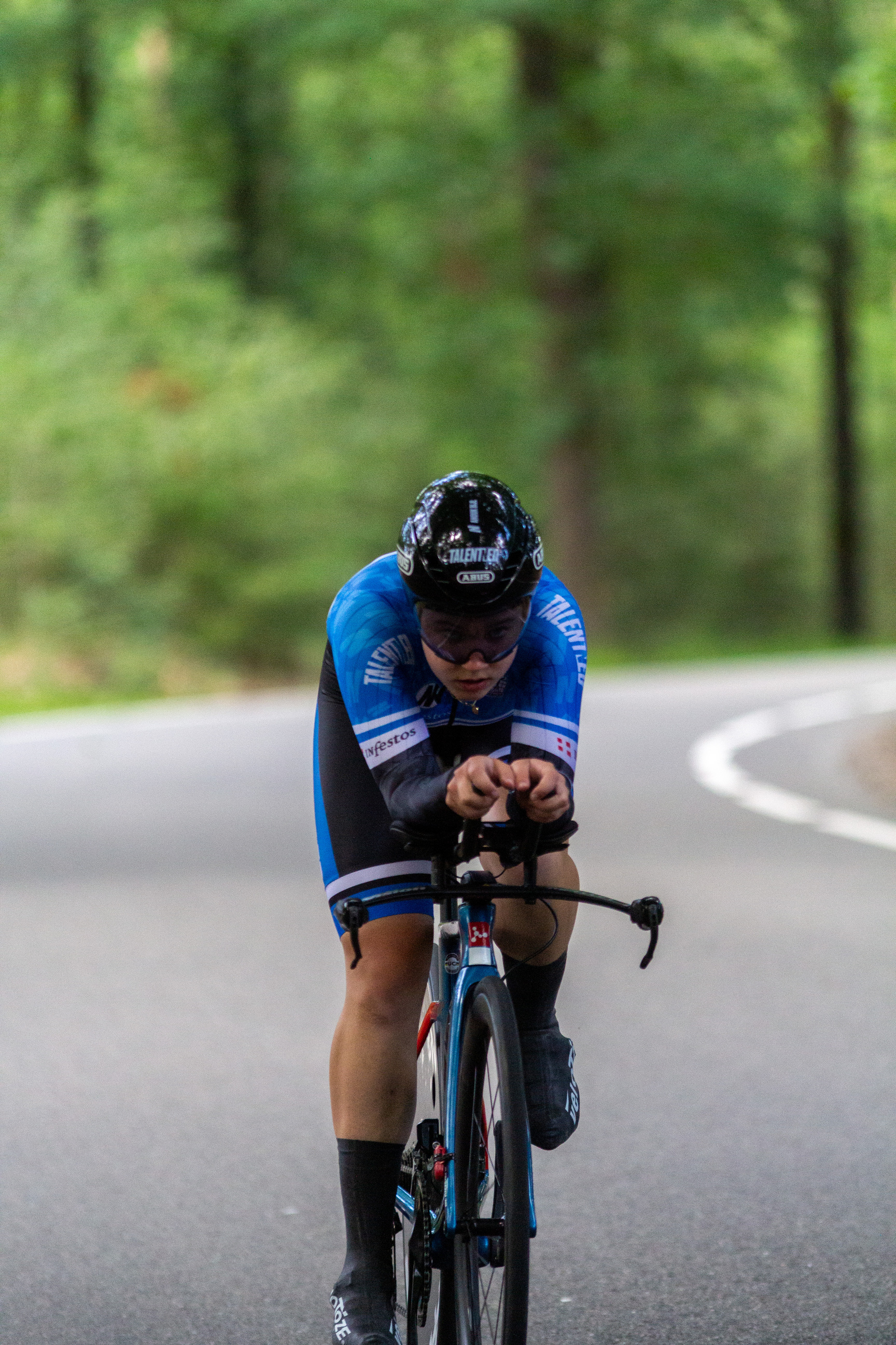 A man wearing a blue Wielrennen racing suit races on his bike on the road.