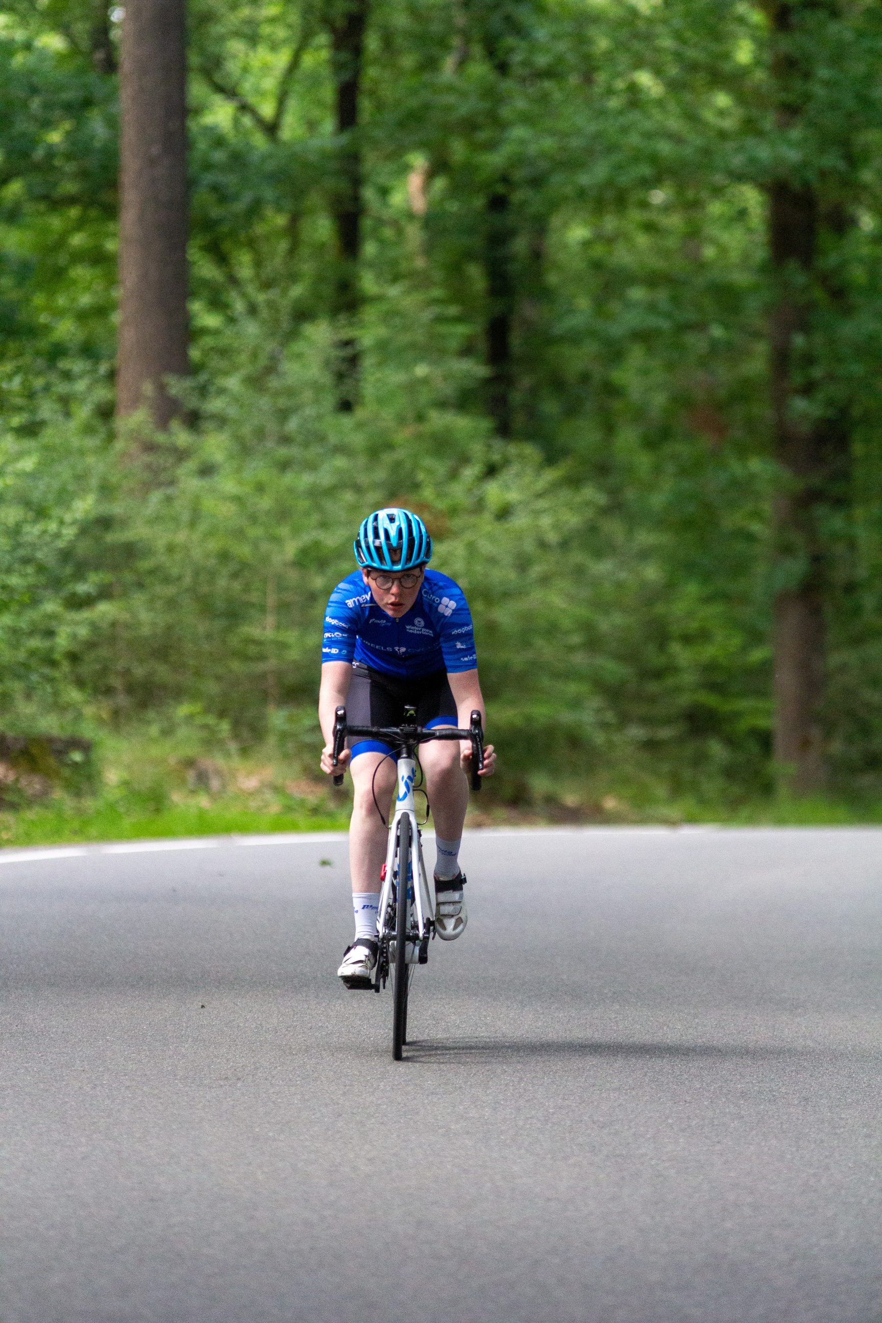 A man on a bike wearing a blue helmet and jersey. He is riding the bike on a road surrounded by trees.