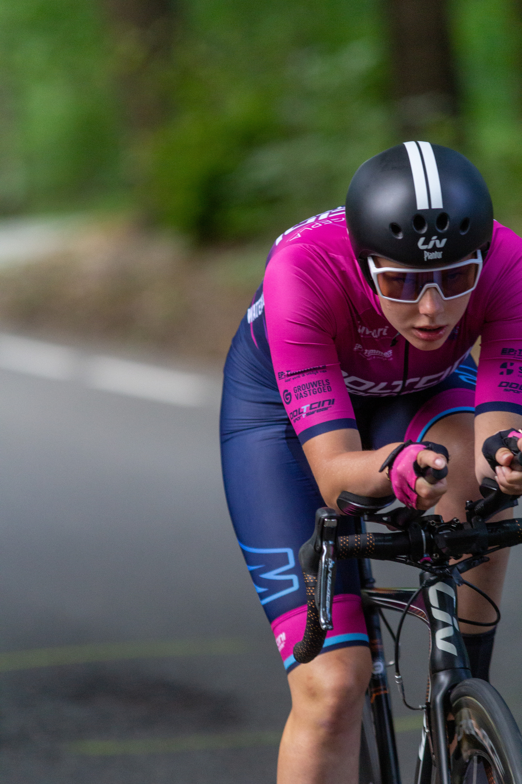 A young woman is riding a bicycle in a race. She has a black helmet on and a purple jersey with the name "Wielrennen".